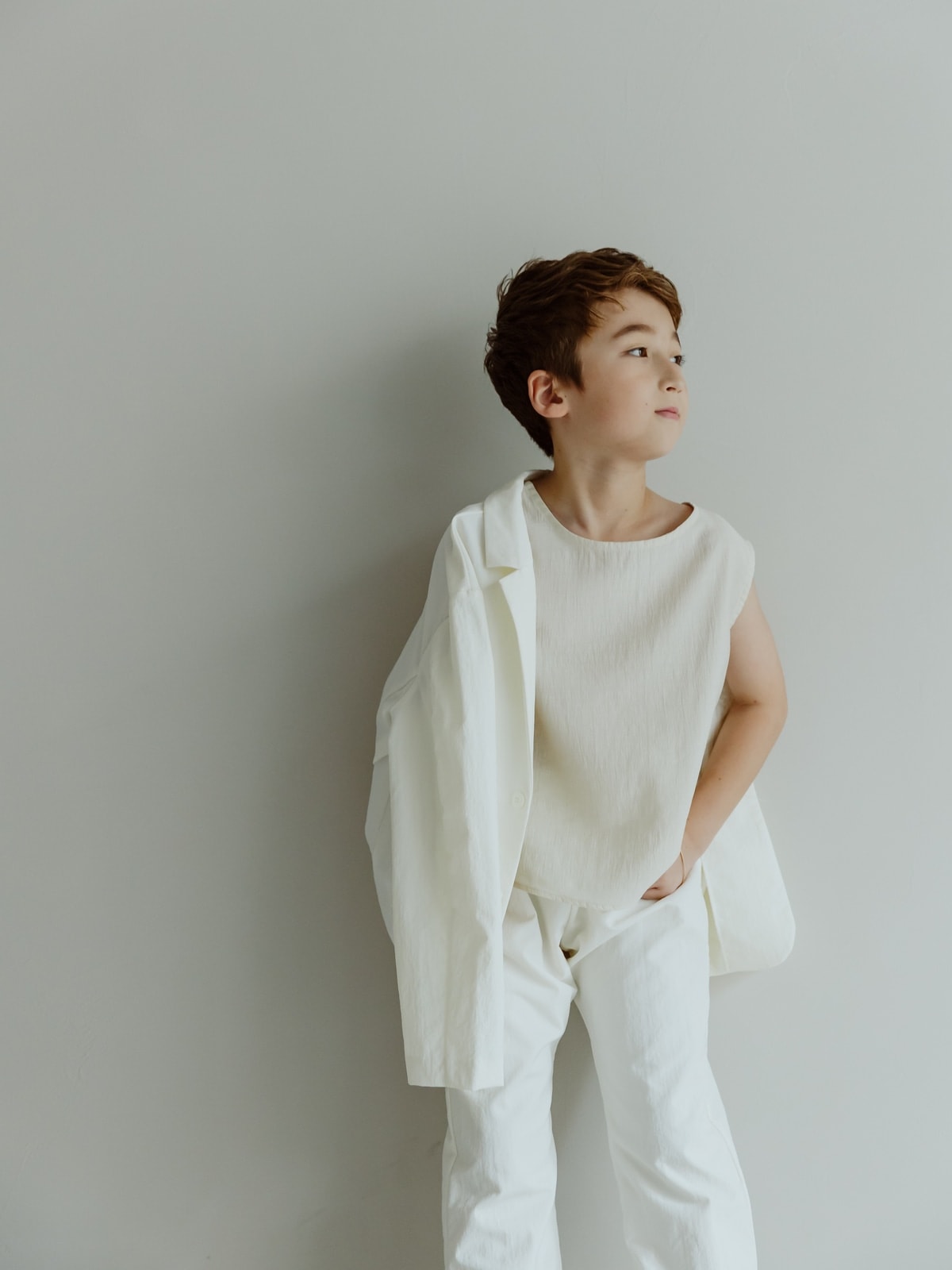 白いノースリーブのトップスと白いパンツを着た少年が、白いシャツを片肩に羽織り、淡いグレーの壁を背景に斜め右上を見つめる全身ポートレート。