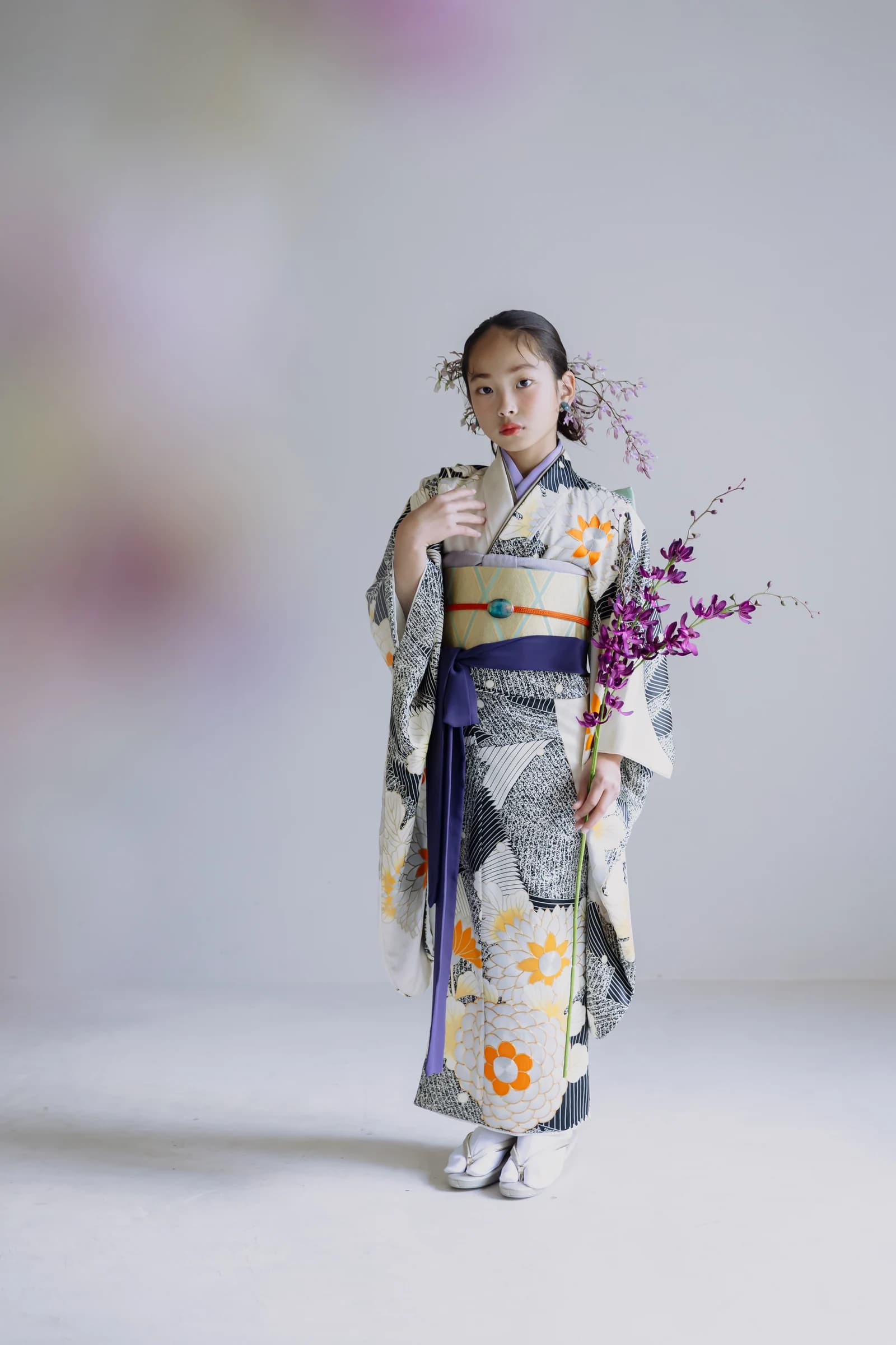白地に黒と橙の大輪花柄の振袖を着た少女が紫色の蘭の枝を手に立つ全身像。黄の帯に紫の帯締めを合わせ、淡い背景の正面構図。
