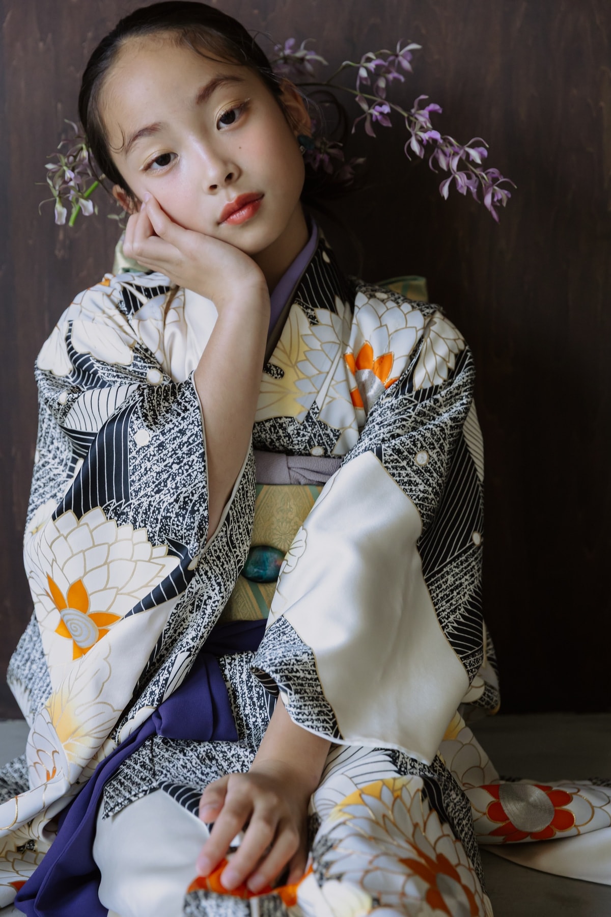 白地に黒い縞と橙・白の花柄の着物・紫の袴姿の少女が、紫の蘭の花を背景に頬杖をついて座る縦位置のクローズアップ。
