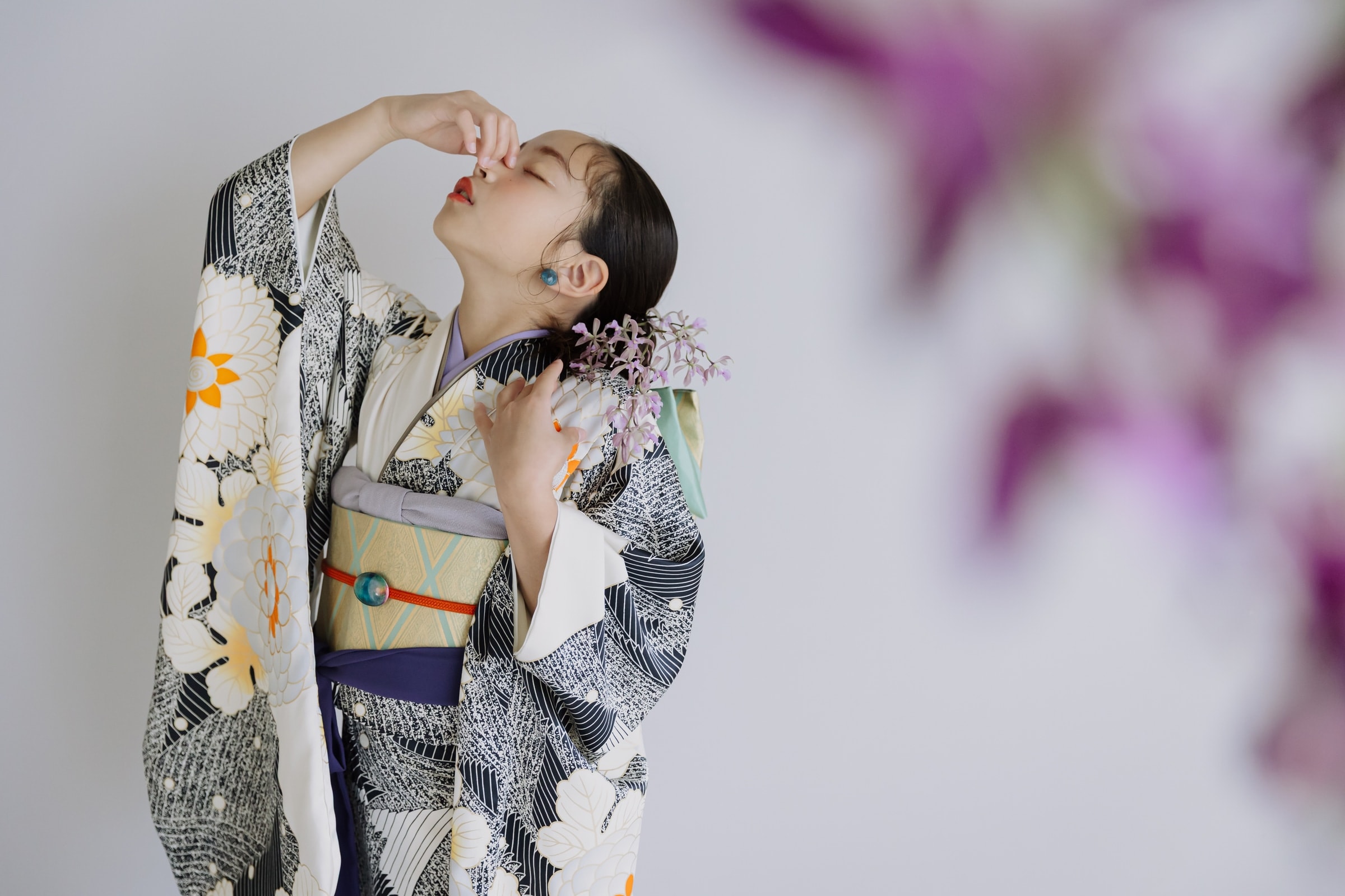 白地に黒い縞と橙・白の花柄の着物に金色の帯を合わせた女性が、生花の髪飾りを付け、片手を顔に添えて顎を上げる立ち姿。手前に紫の蘭の花がぼかしで重なる横位置の構図。