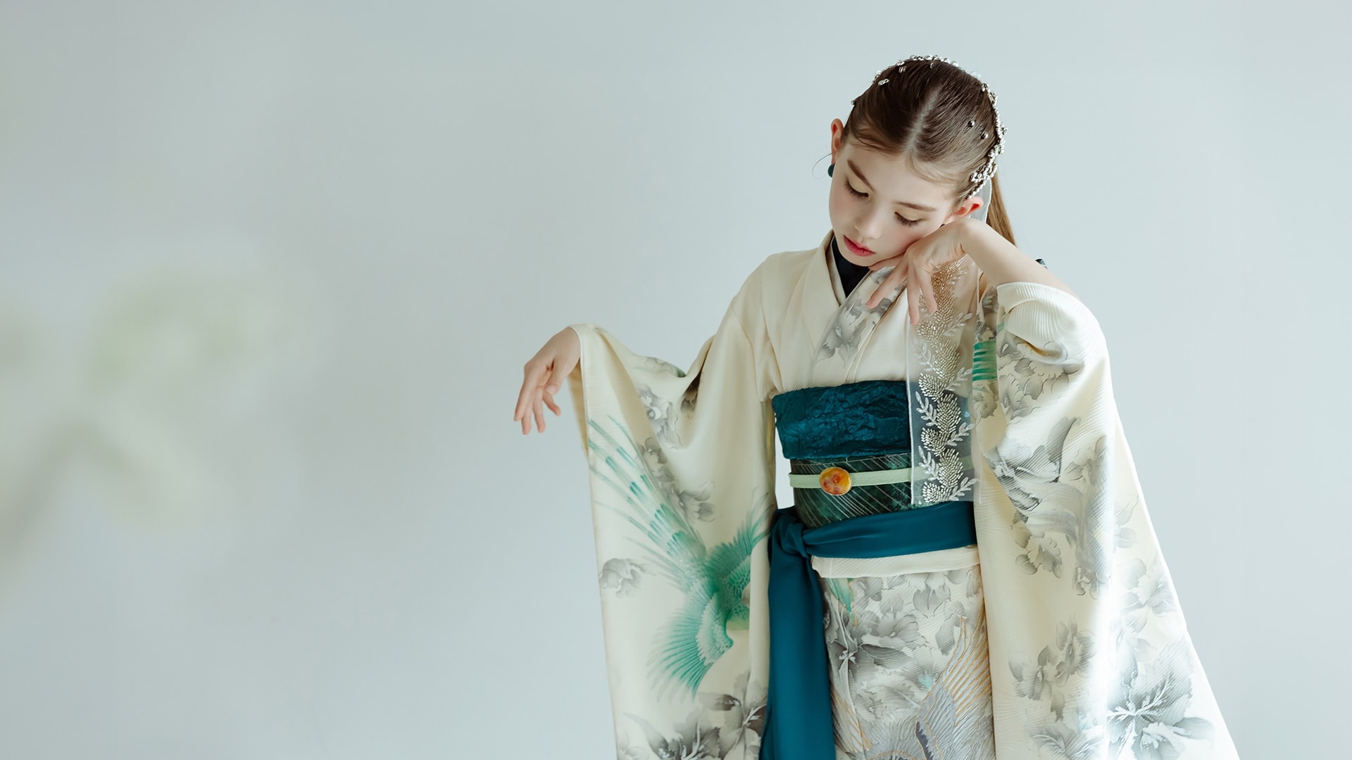 ULUM 着物コレクション — クリーム色地に緑の鳥と花柄の振袖を着た少女が、緑の帯を締め視線を伏せて立つ姿。髪飾りを付け右側に寄った構図、背景は淡いグレー。