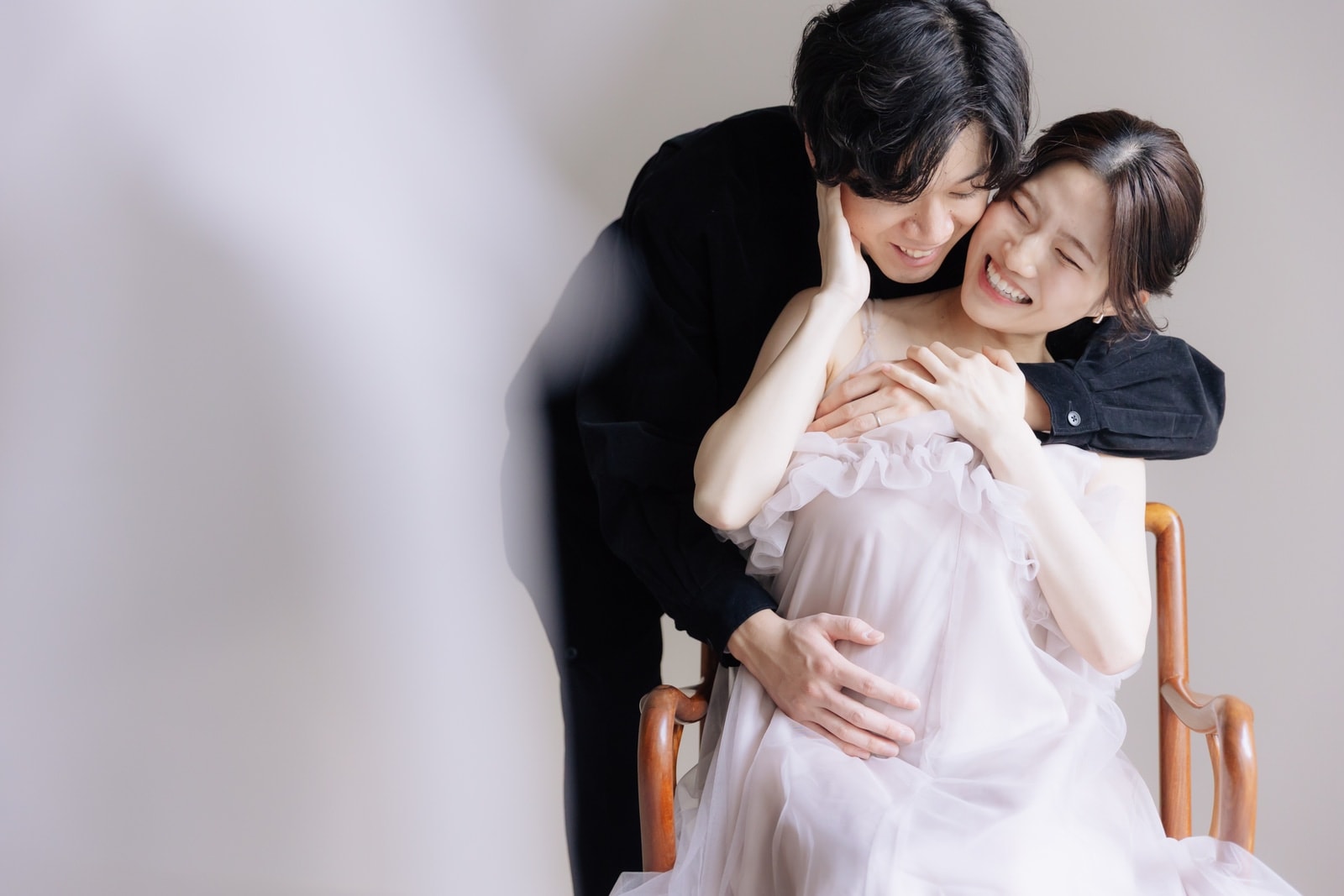 淡いピンクのチュールドレスを着て木製椅子に座る笑顔の女性と、黒い衣装で背後から寄り添う男性の二人。明るい背景の右寄り構図。