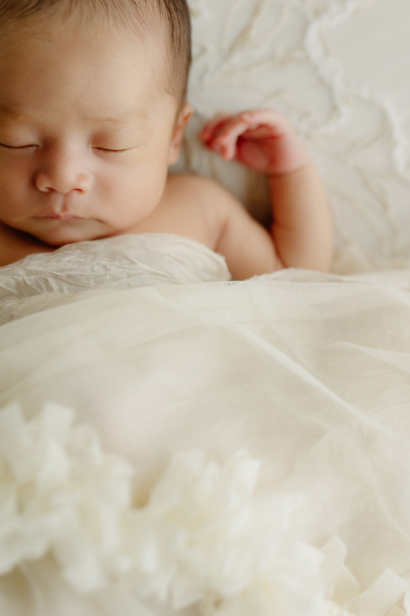 眠る新生児 — クリーム色の薄手の布に包まれ、顔を左に向けて目を閉じ、片手を上げた姿を上半身中心に捉えた接写。前景に白いチュールが広がる構図。
