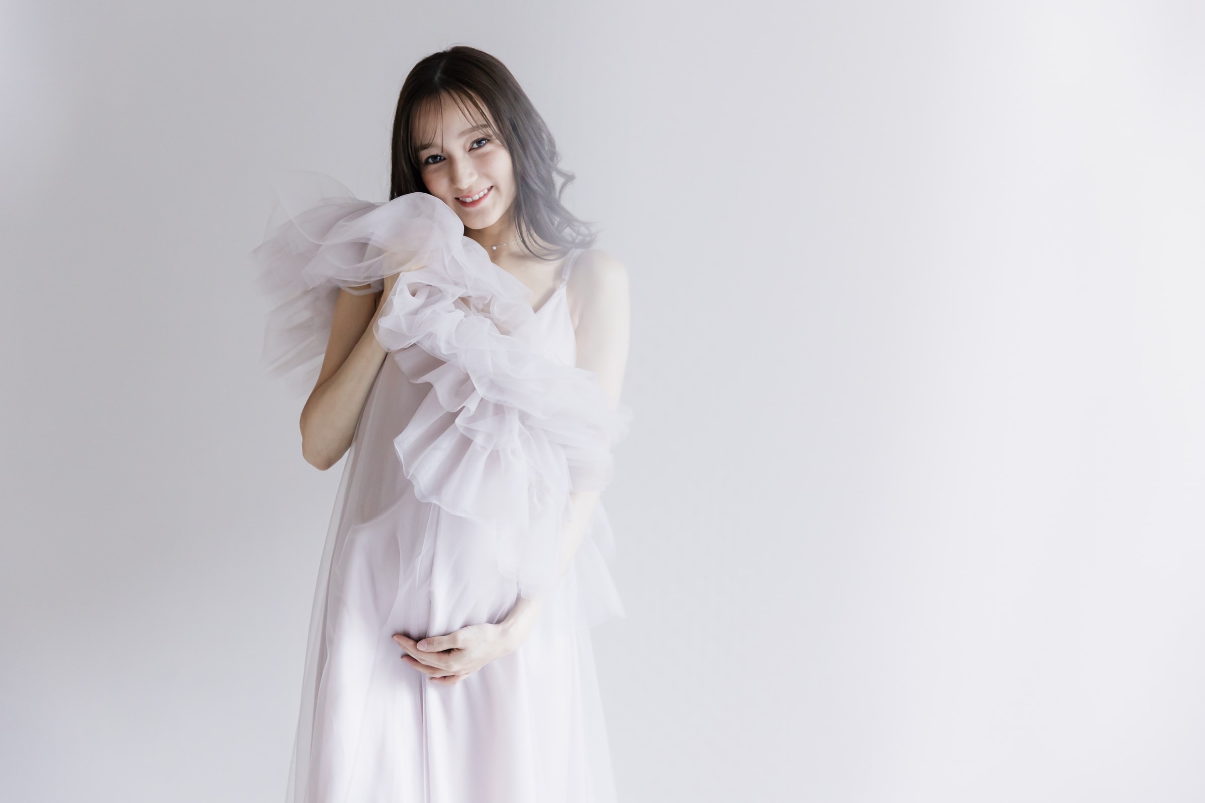 妊娠中の女性 — 淡いラベンダー色のチュール装飾付きドレスを着てお腹に手を添え、白い背景の前で微笑む全身ポートレート。