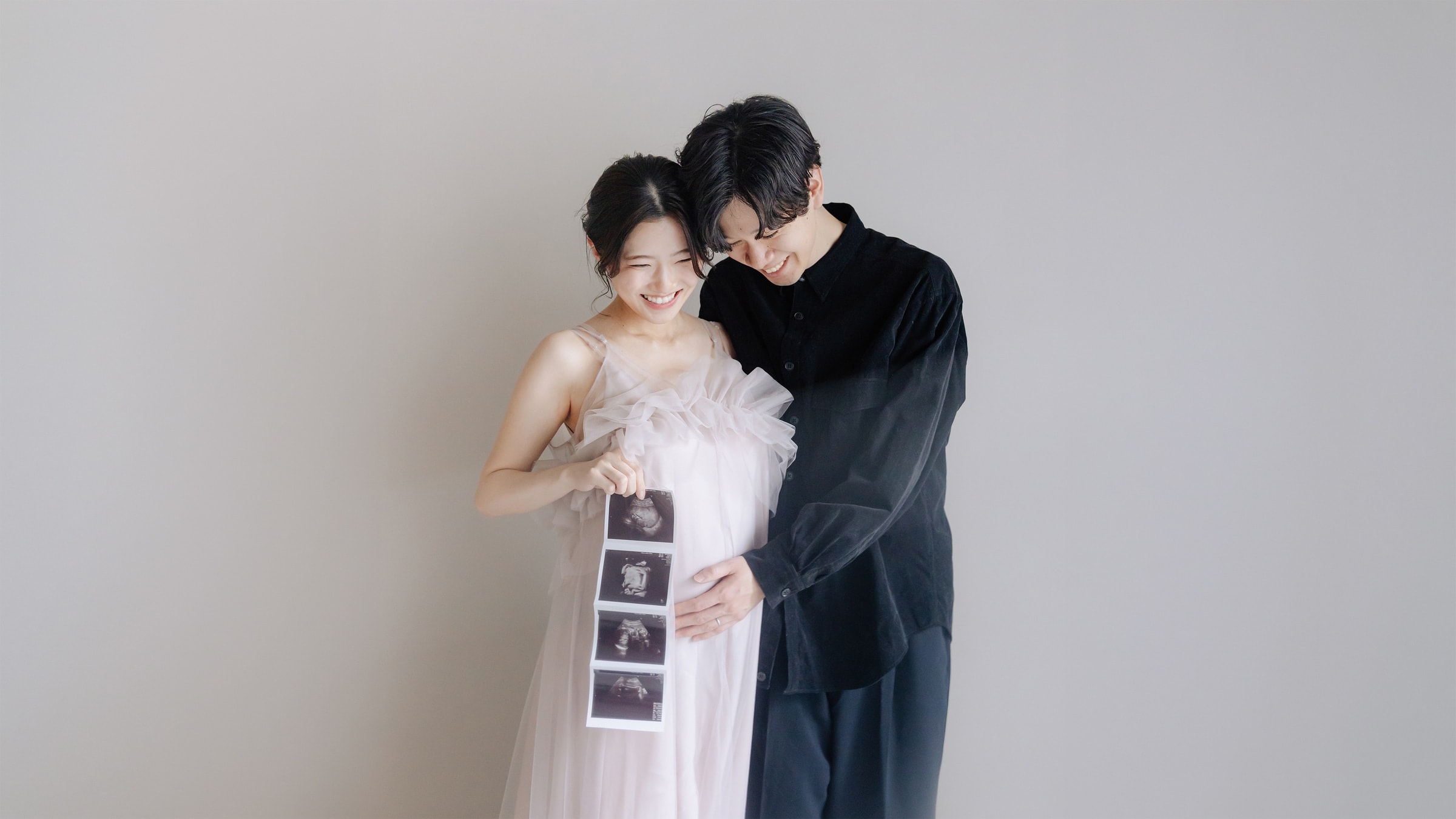 淡いピンクのチュールドレスの妊婦と黒シャツの男性が寄り添い、エコー写真を持って微笑むマタニティ撮影。灰色背景の中央に二人が立つ構図。