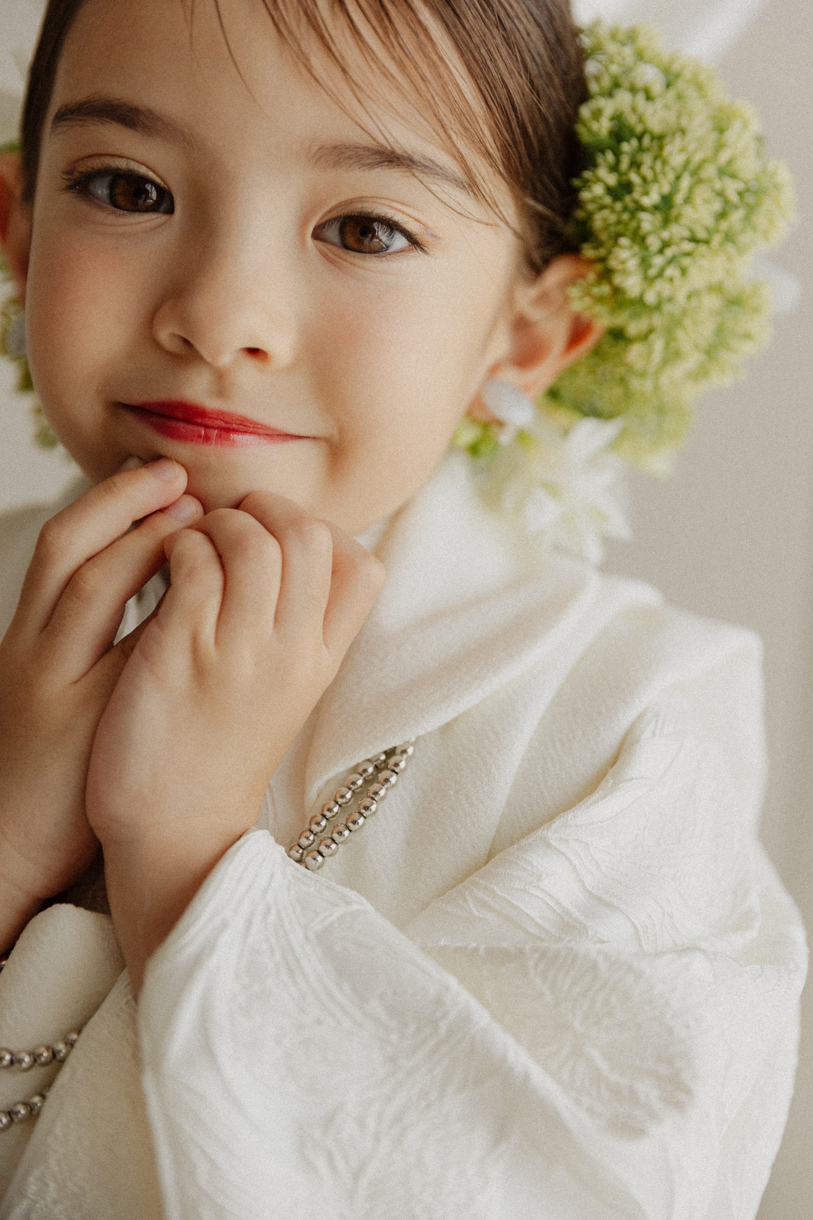 白い地紋入りの着物を着た子ども — 髪に淡緑の花飾りをあしらい、両手を口元に添えた上半身のクローズアップ、明るい背景。