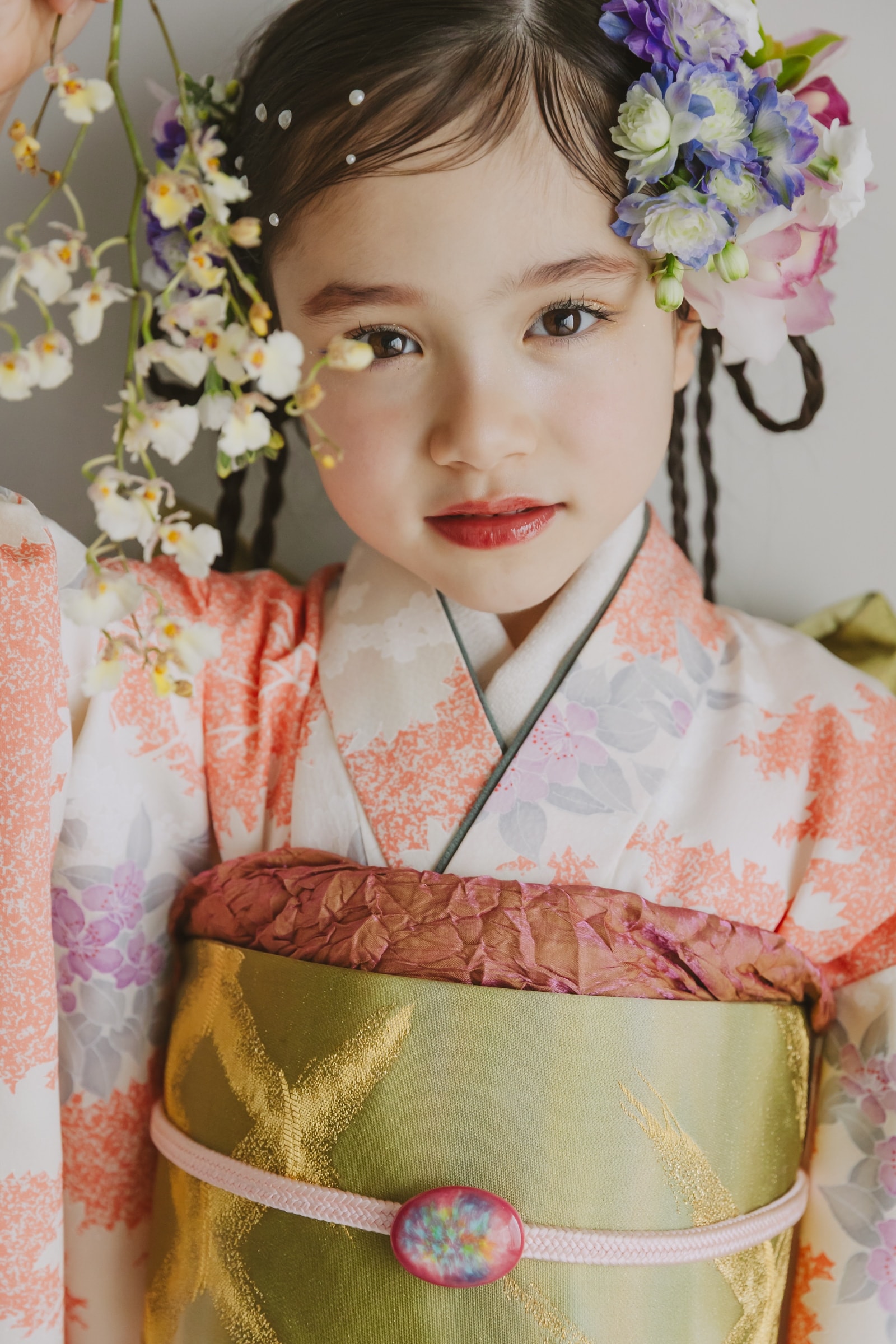 白地に橙と桜花柄の着物を着た女児の上半身。金色の帯と桜色の帯締め、紫や白の花の髪飾りを付け、正面を向く。背景は淡い灰色。