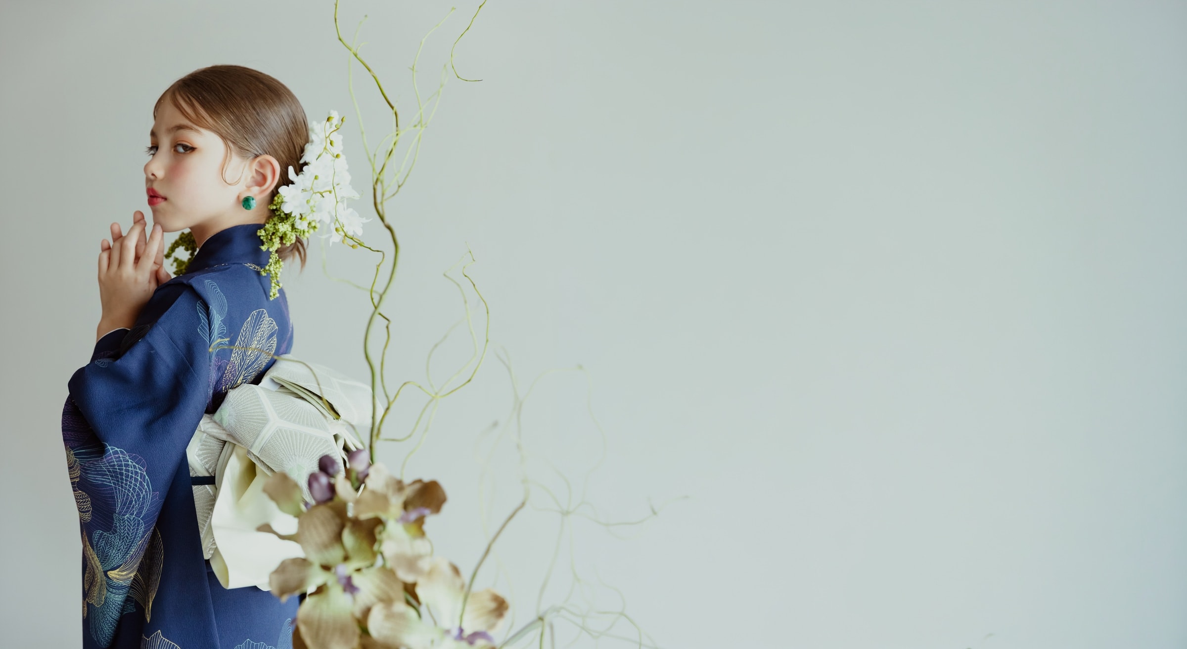 紺色地に線描の花柄を配した着物姿の少女が横向きで立ち、白い帯を結び髪に白い花飾りを添える。手前に枯れた花と曲がった枝、背景は淡い灰緑で右側に余白。