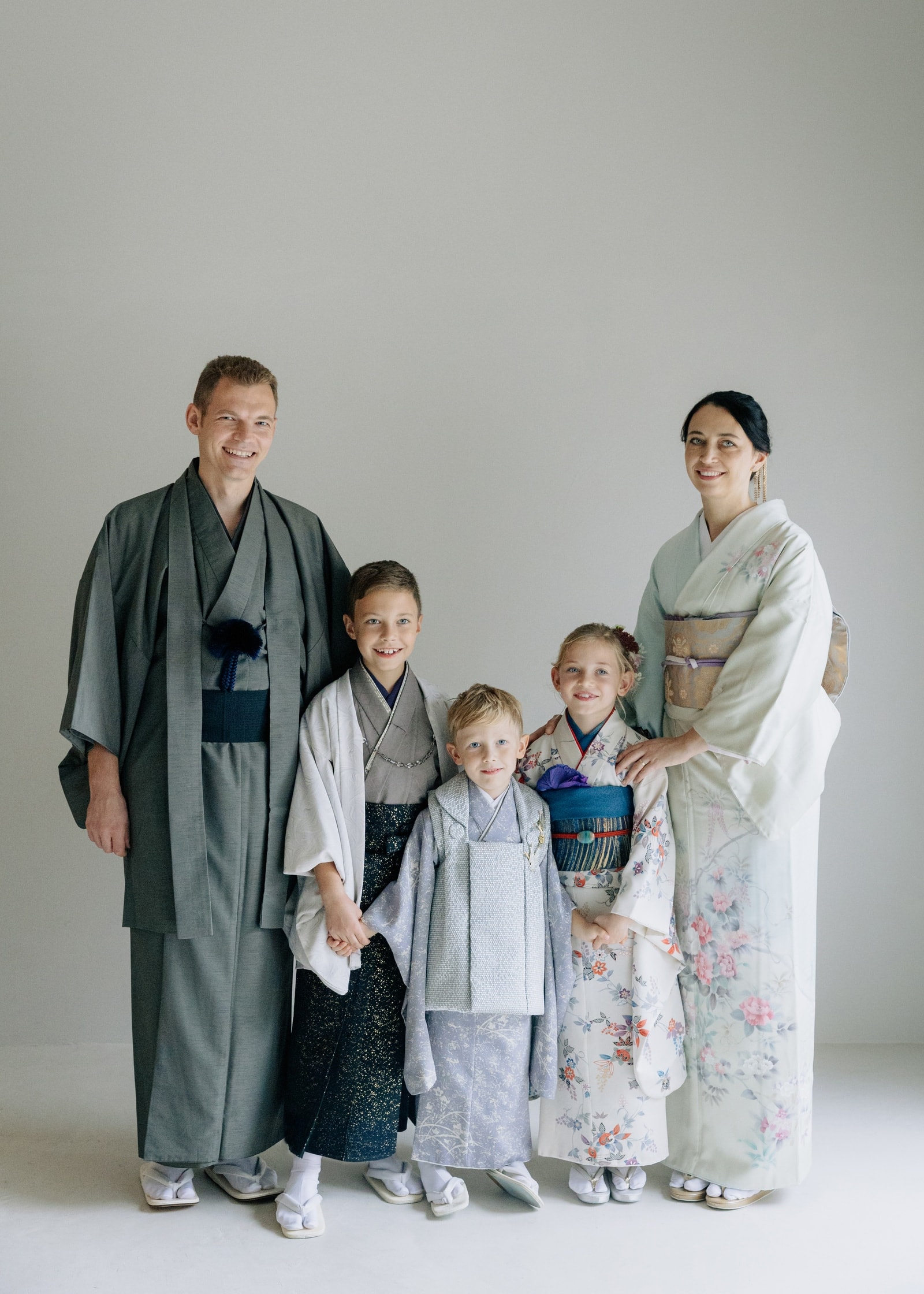 和装の家族5人が並ぶ全身写真。男性は灰色の着物と袴、女性は花柄の薄緑色の着物、子ども3人は灰色や水色、花柄の着物を着用、無地の背景。
