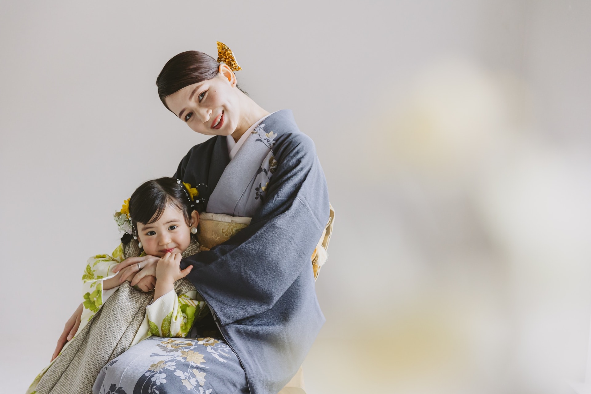 青灰色に花柄をあしらった着物の女性と、緑と白の花柄の着物を着た女児が寄り添う構図。背景は淡い無地で、二人とも左寄せに配置され微笑んでいる。