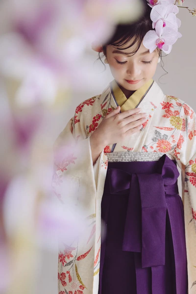 クリーム地に赤や橙の花柄をあしらった着物に濃紫の袴を合わせた女性 — 髪に蘭の花を飾り胸に手を添え目を伏せる、手前に淡い花がぼけて入る縦長構図。