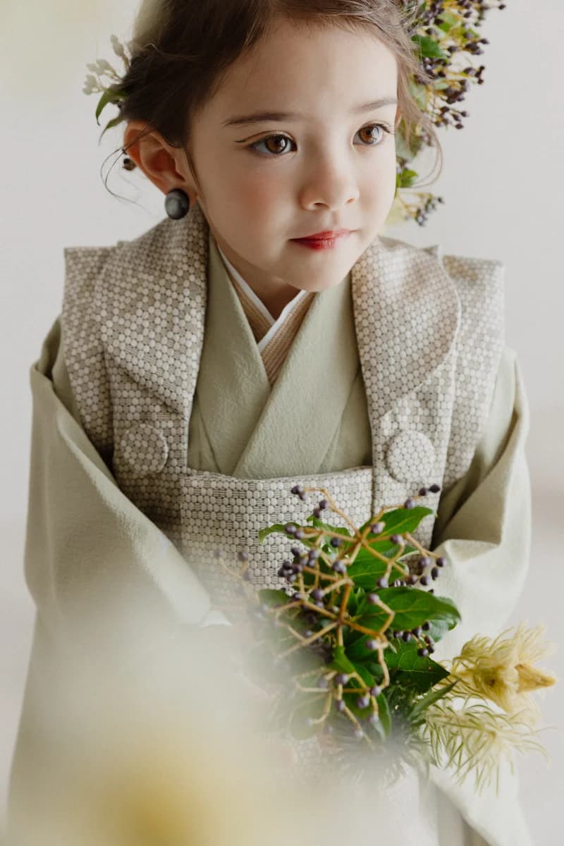 淡い緑色の被布を重ねた七五三の女の子。白い水玉模様の被布と着物姿で、髪に小花や実物の飾りを添え、紫の実と緑葉の小物を手にした上半身の縦長構図。