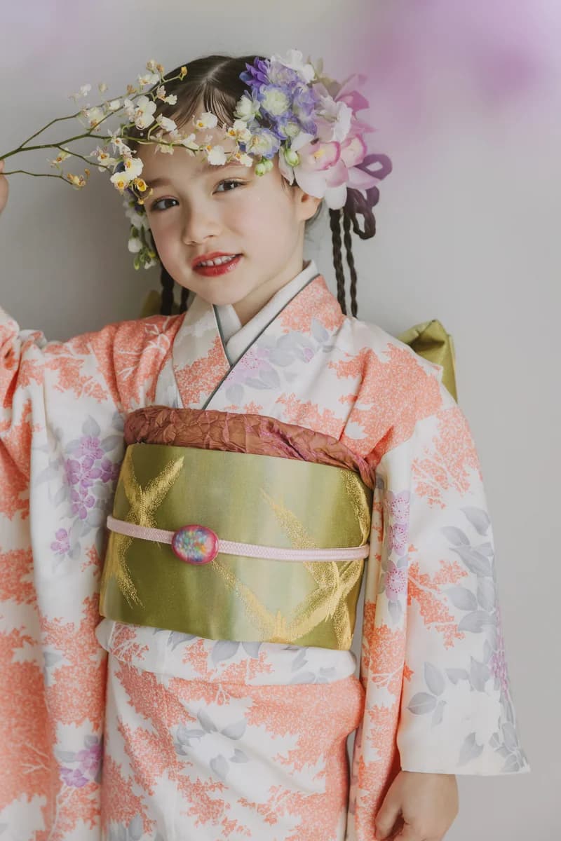 白地にオレンジと紫の草花を散らした着物を着た女児 — 黄緑の帯に紫やピンクの花飾りを頭に飾り、枝を手に上半身を写した縦長構図