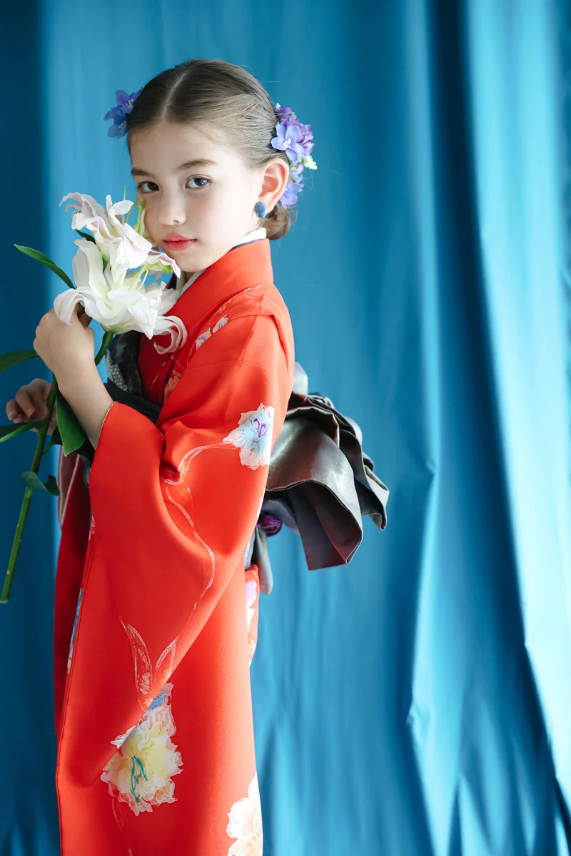 赤地に白や黄の花柄を配した七五三着物姿の女児 — 紫の花の髪飾りを付け白い花を持ち、青いドレープ背景の前で横向きに立つ縦長構図。