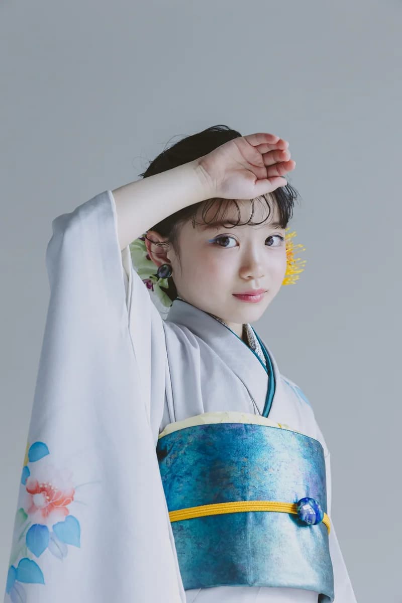 白地に水色と橙の花柄の着物に青緑の帯と黄色帯締めを合わせた女の子が、片手を額にかざし横を向く上半身の縦構図。黄色の髪飾りを付けている。