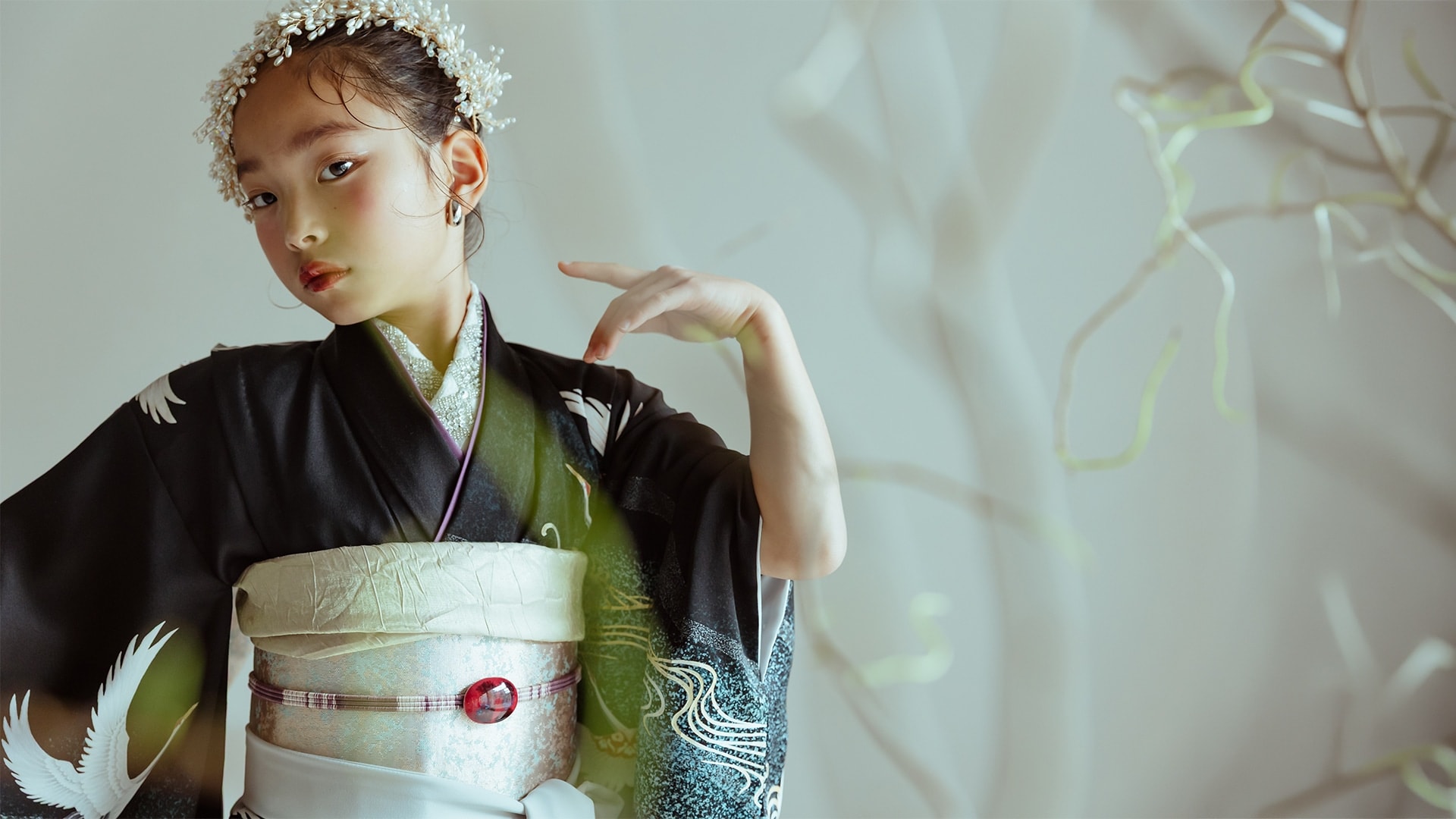 黒地に鶴と緑のぼかし柄の着物を着た少女 — 銀色の帯に赤い帯留め、白い花飾りを頭に付け、片手を顔の横に上げた半身の構図。背景は枝のシルエット。