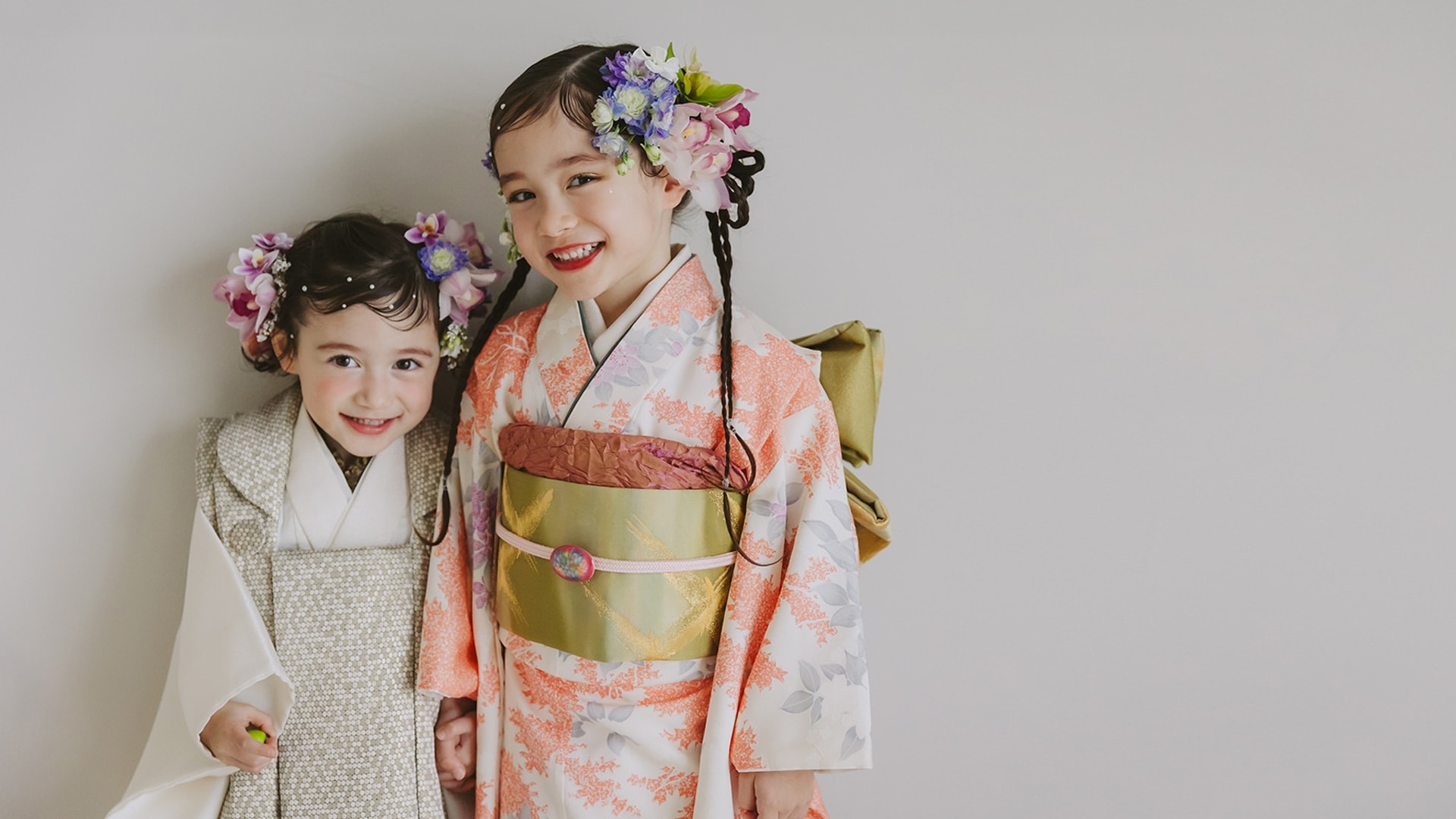 七五三の和装をした二人の子ども — 右の子は白地に橙色の草花柄の着物と黄緑の帯、左の子は白地の被布、ともに花飾りを付け灰色背景の前で並ぶ。