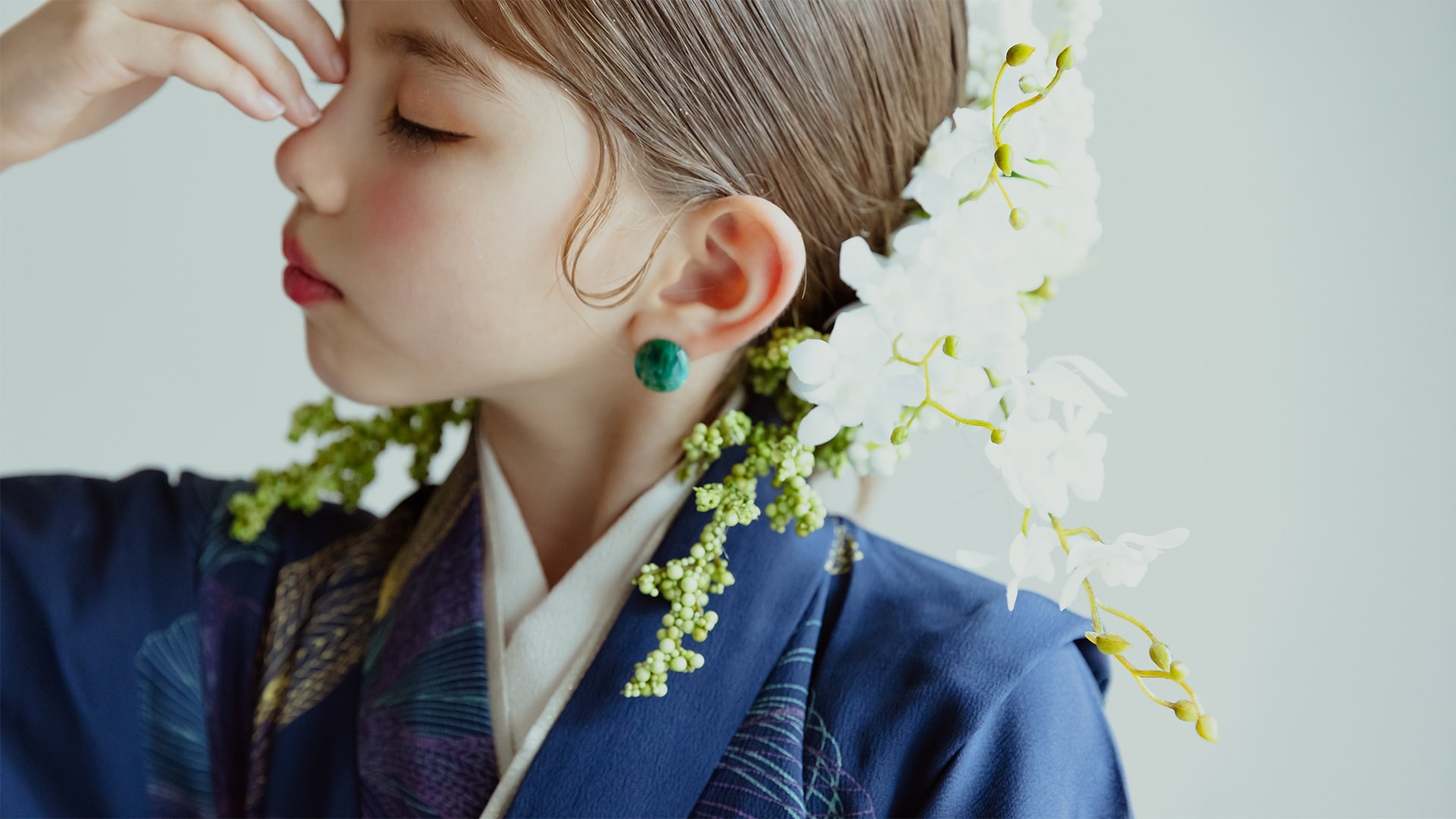 紺色地に紫や金の柄をあしらった着物姿の子どもの横顔 — 緑の丸い耳飾りを着け、白い蘭と緑の小花の髪飾りを添えた構図。
