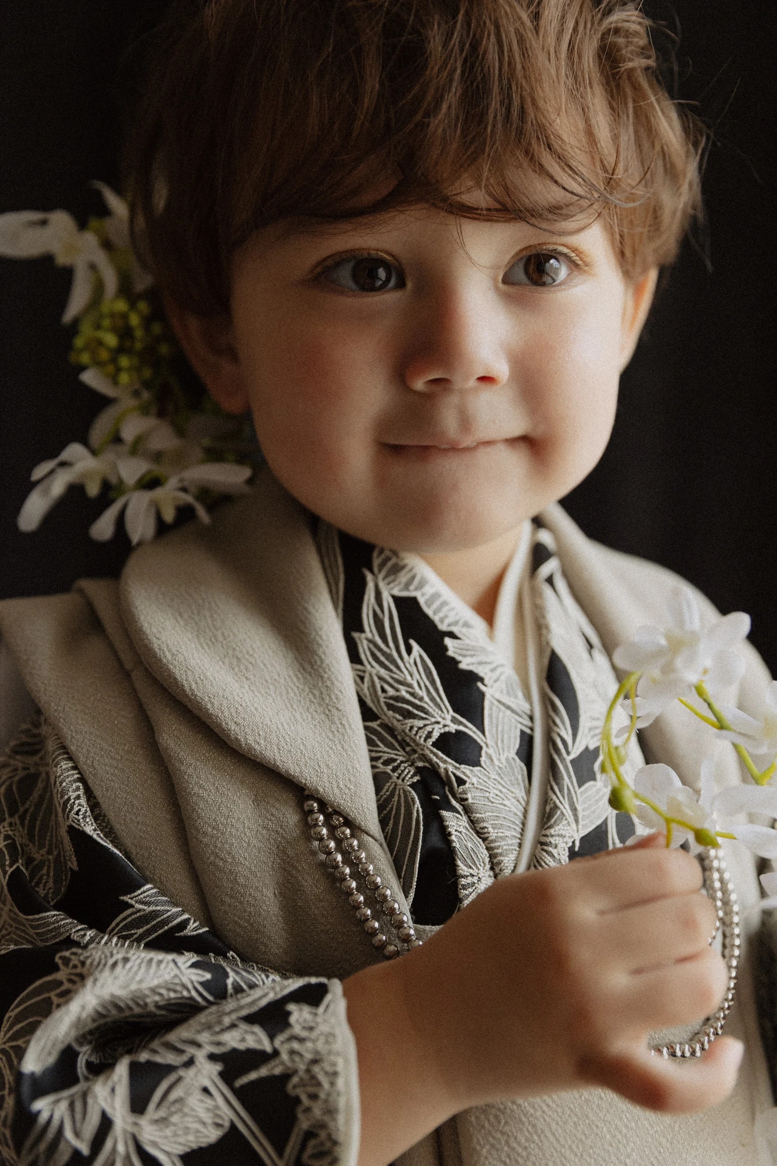 黒地に白の草花柄の着物にベージュの羽織を重ねた幼児の上半身。白い花と銀色のビーズを手にし、髪に白い花飾りを添えて正面を見るクローズアップ構図。