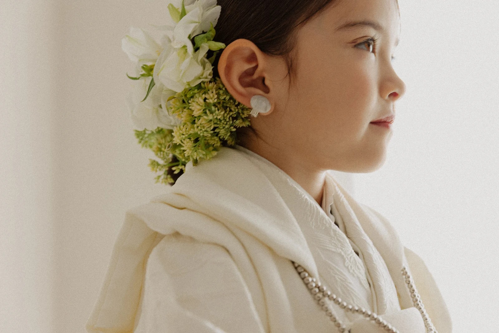 白い祝着姿の幼児を右向きの横顔で捉えた接写。耳元に白い花と緑の小花の髪飾り、白い丸いイヤリングとパール調の首飾りを添えた構図。背景は淡い白。