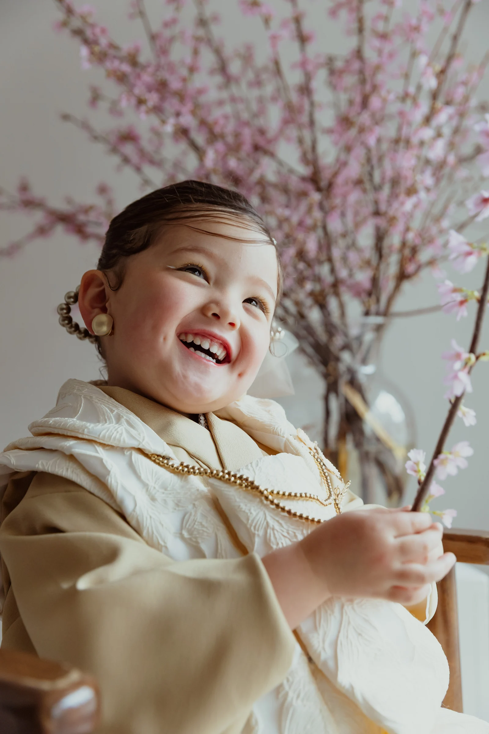 生成り色の被布と着物を着た幼児が桜の枝を手に笑顔を見せる。金色のビーズ飾りと丸い耳飾りを添え、背景にピンクの桜が広がる縦構図のポートレート。