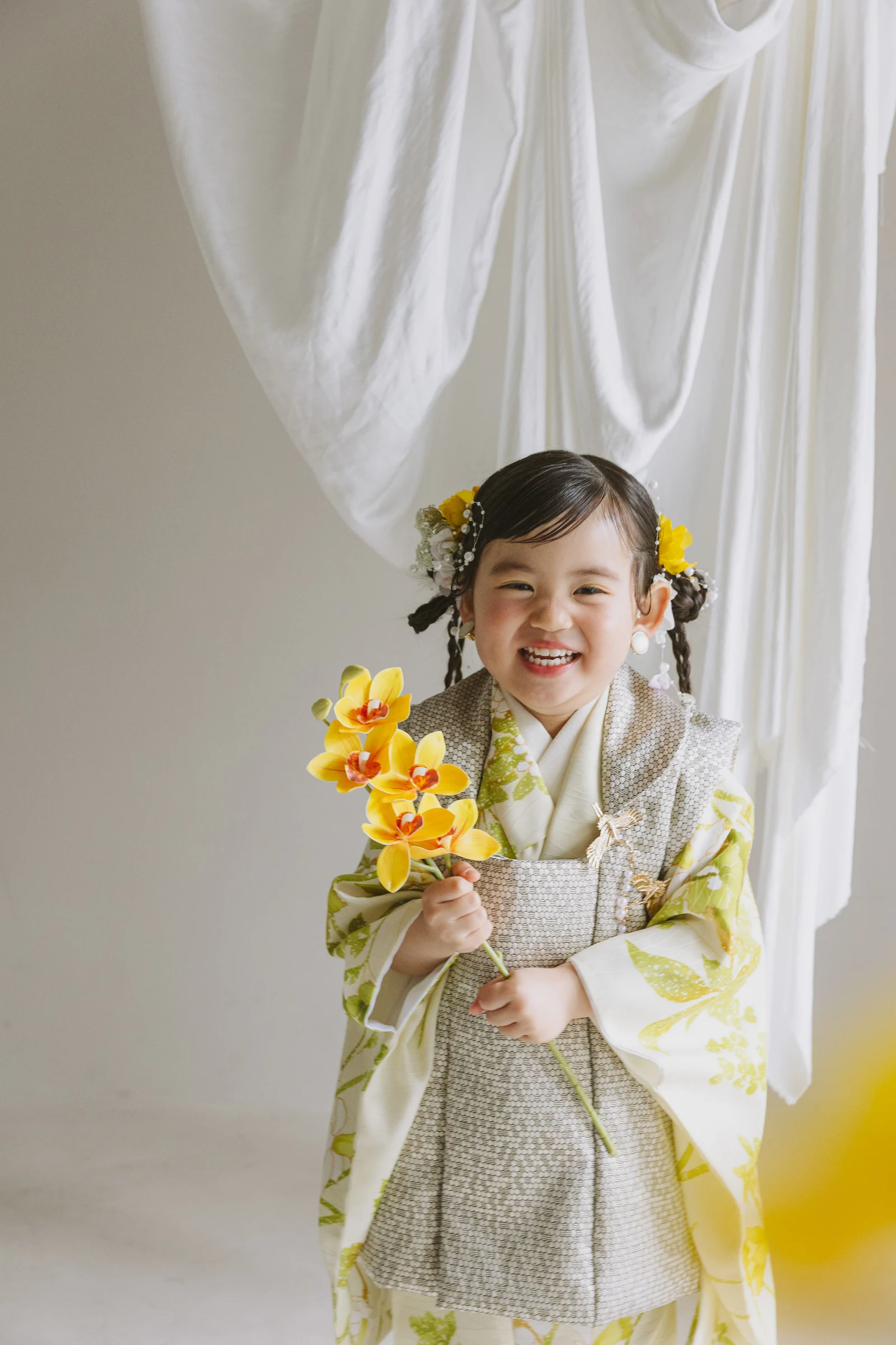黄緑の草花柄の着物にグレー被布を重ねた3歳児が、黄色い蘭の花を手に笑顔で立つ。白い布を背景にした正面構図のポートレート。