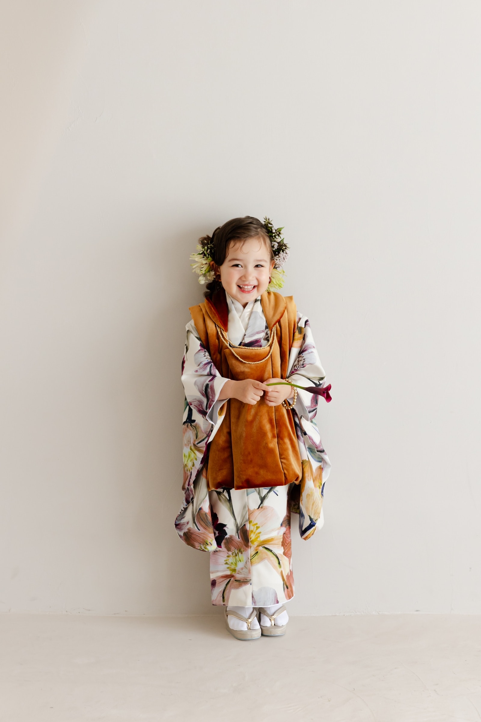 白地に水彩調の花柄の着物とキャメル色のベロア被布を着た女児が、紫のカラーの花を胸元に持ち満面の笑みで正面に立つ全身像。背景は淡い灰色の壁。