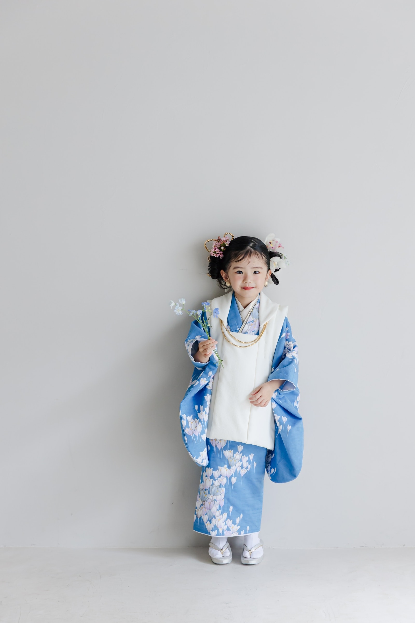 青地に白い花柄の着物に白い被布を重ねた七五三姿の女児が、髪飾りを付け青い小花を手に、白い背景の前で正面を向いて立つ全身像。