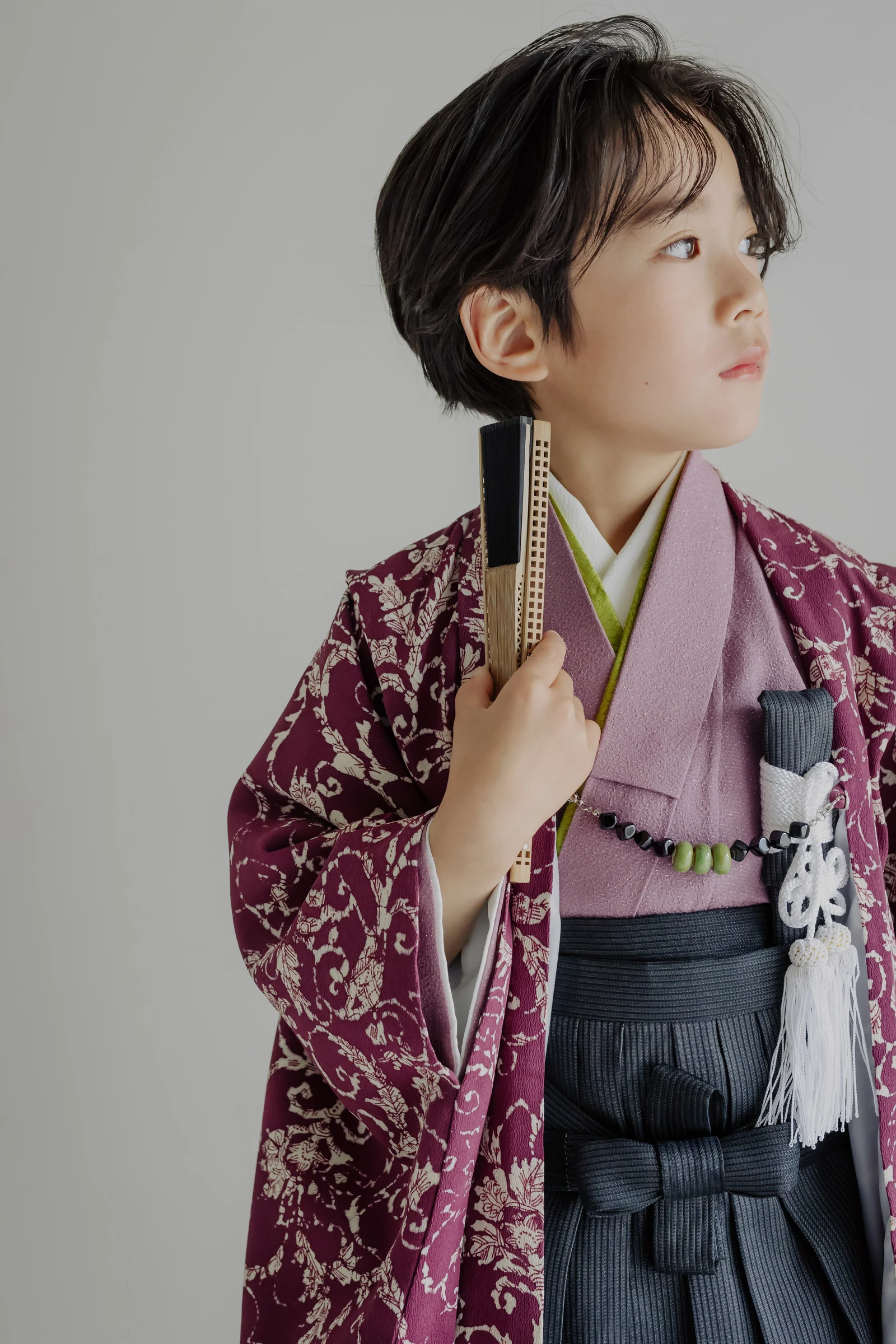 臙脂色に唐草模様の羽織と藤色の着物、灰色の袴をまとった男児が扇子を手に横を向く半身像。白い房飾りを下げ、無地の背景に上半身を写している。