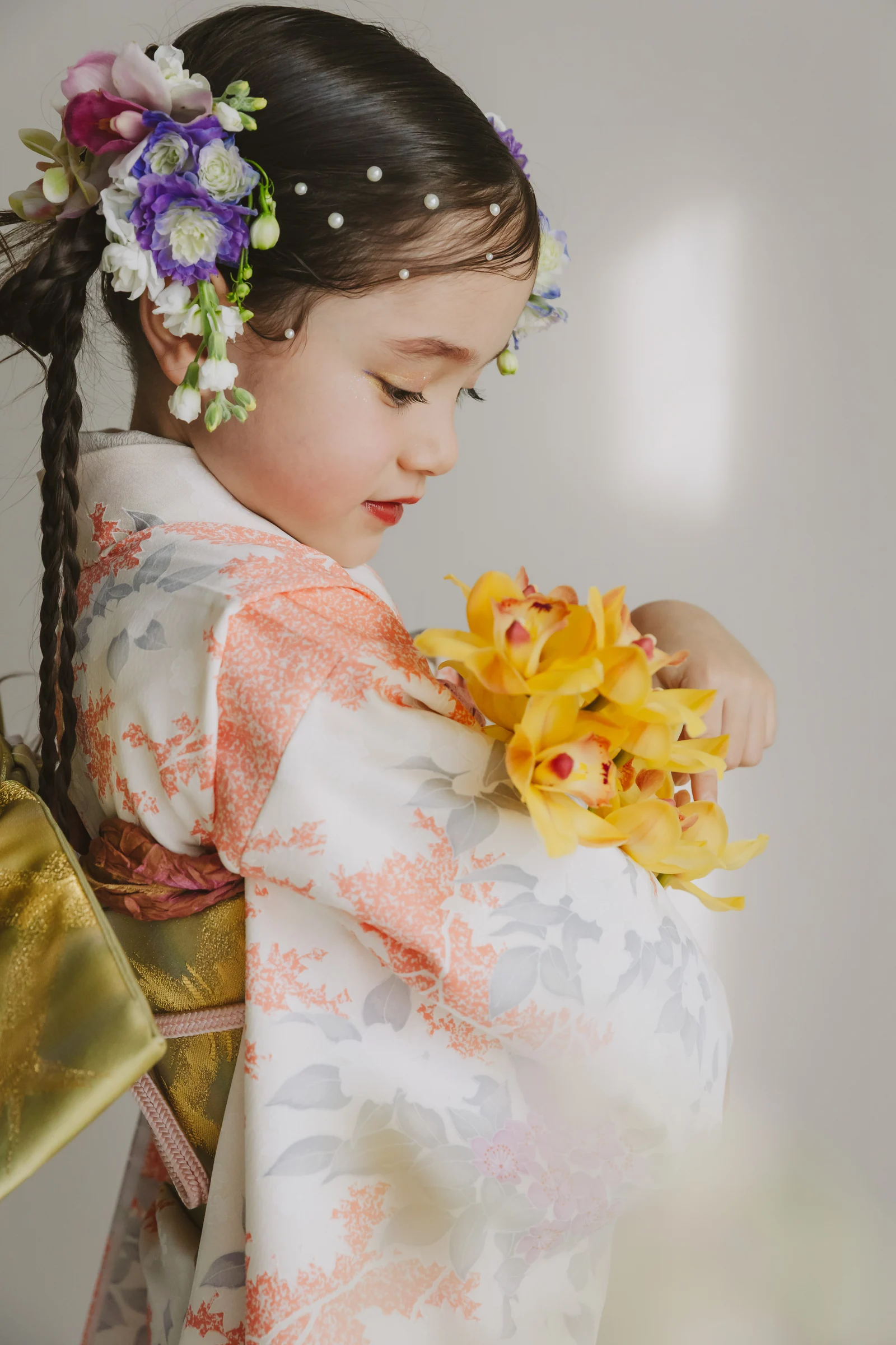 白地に珊瑚色とグレーの花柄の七五三着物を着た女児の横顔。紫と白の花とパールの髪飾り、金色の帯、黄色い花を抱える構図。