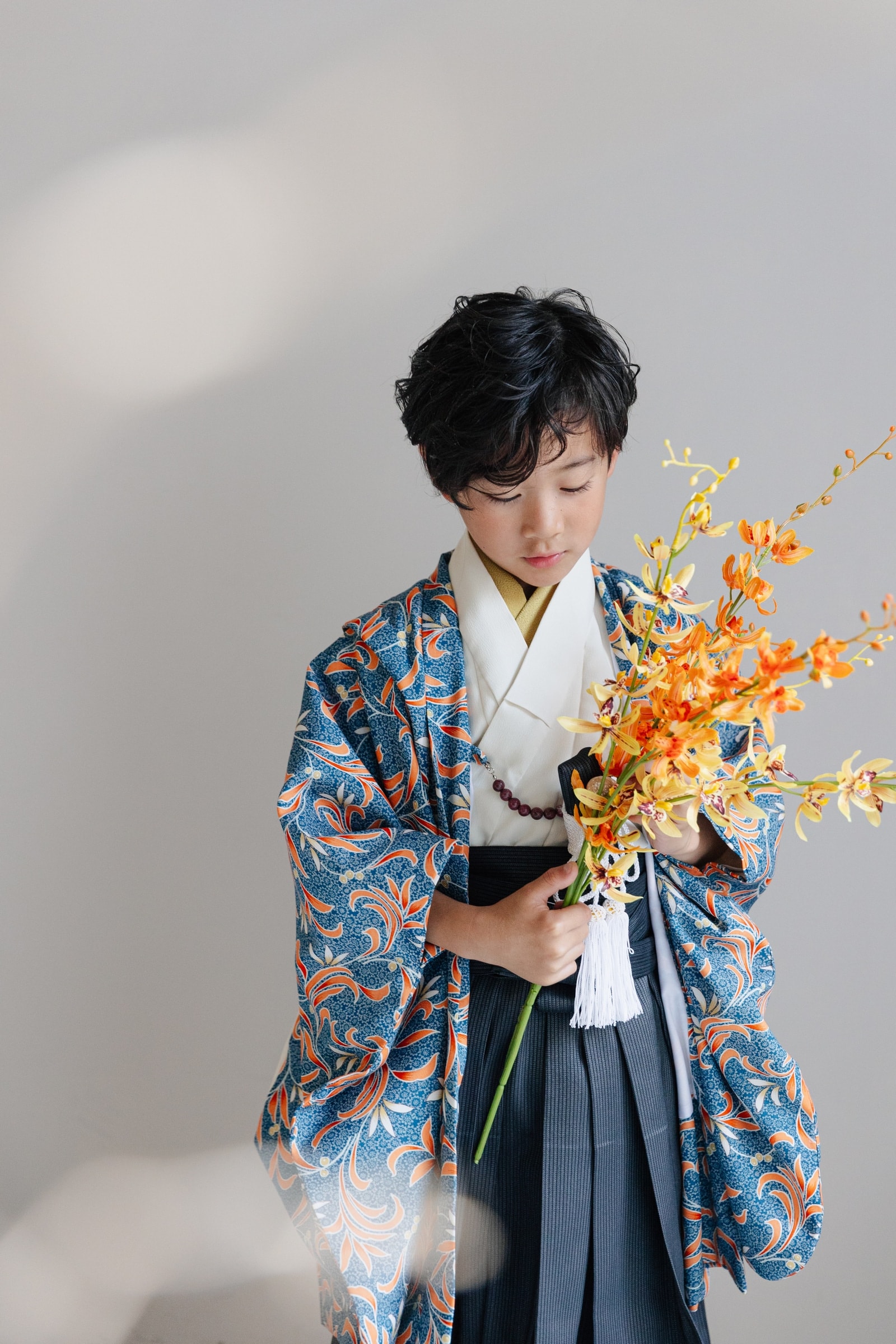 青地に橙の唐草花柄の羽織に縞模様の灰紺袴を合わせた男児が、橙と黄の蘭の花束を手に視線を下げて立つ。背景は淡い灰色、白い半襟と房付き飾り紐が映える。