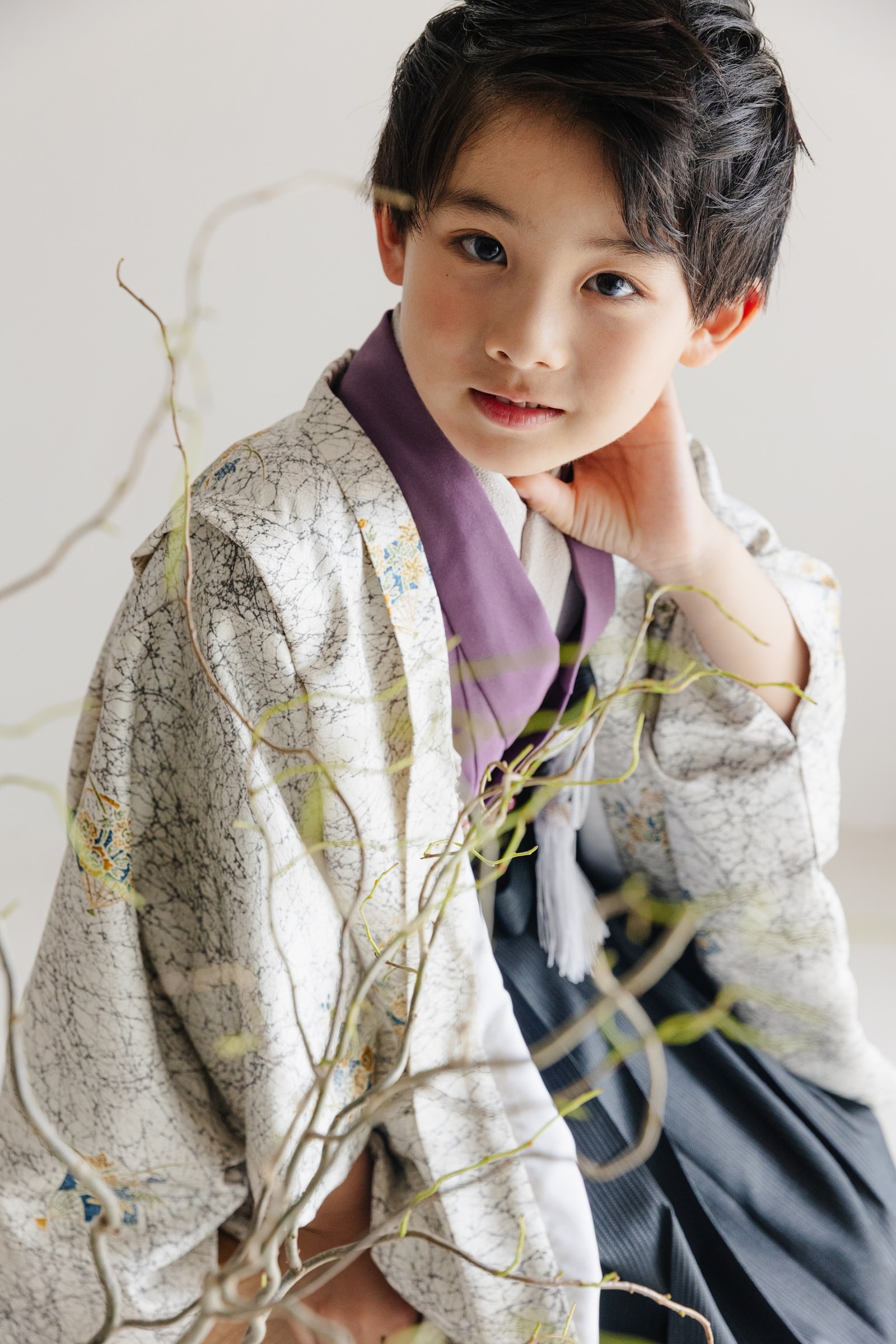 白地に墨流し文様と小花を散らした羽織に紫の襟元、紺の袴を合わせた男児が片手を首に添える上半身の構図。前景に曲がった枝。