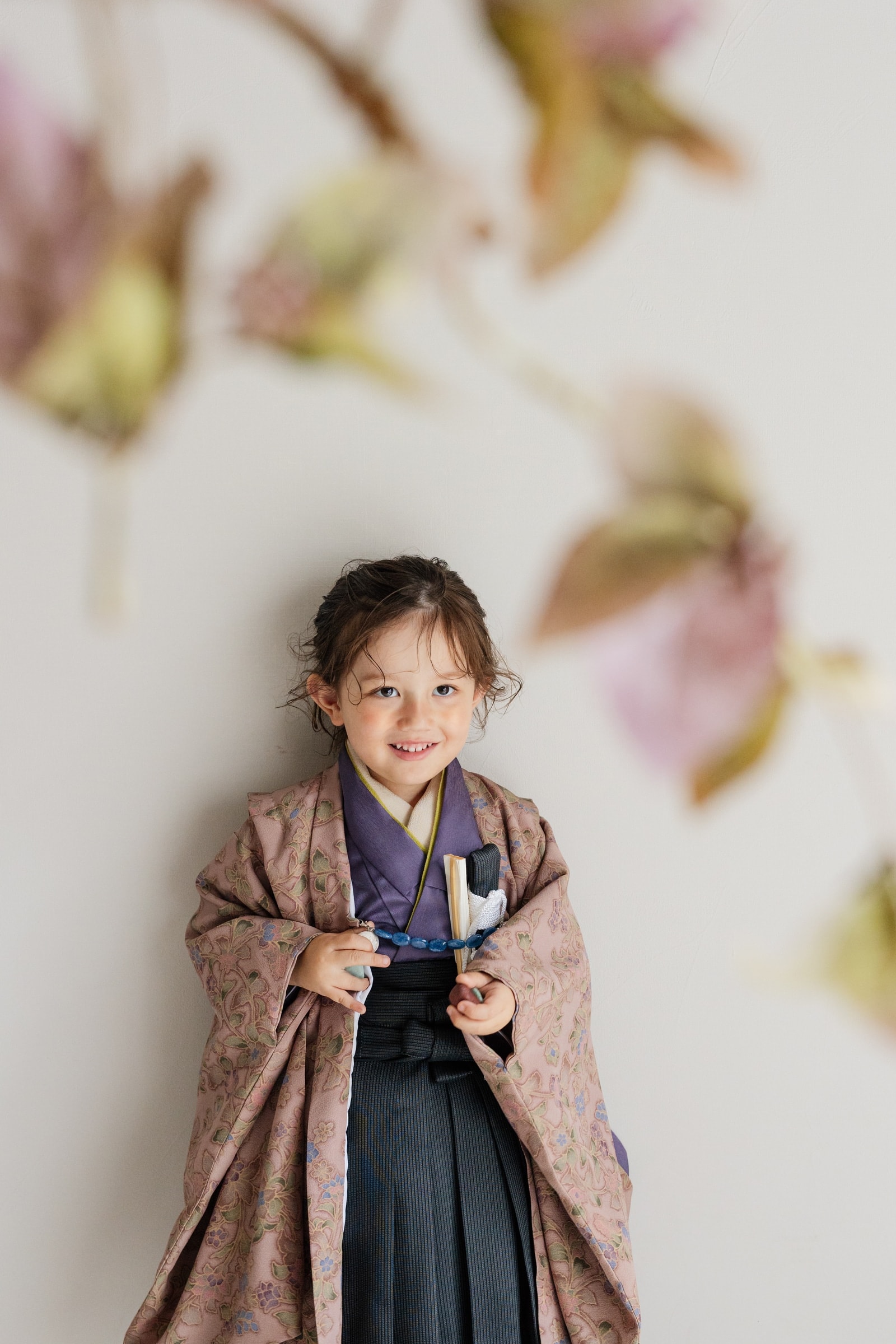 くすみ桃色に草花柄をあしらった羽織を纏う子ども — 紫の着物と黒の袴、扇子と数珠を手に、ぼかした花枝越しの構図で微笑む正面ポートレート。