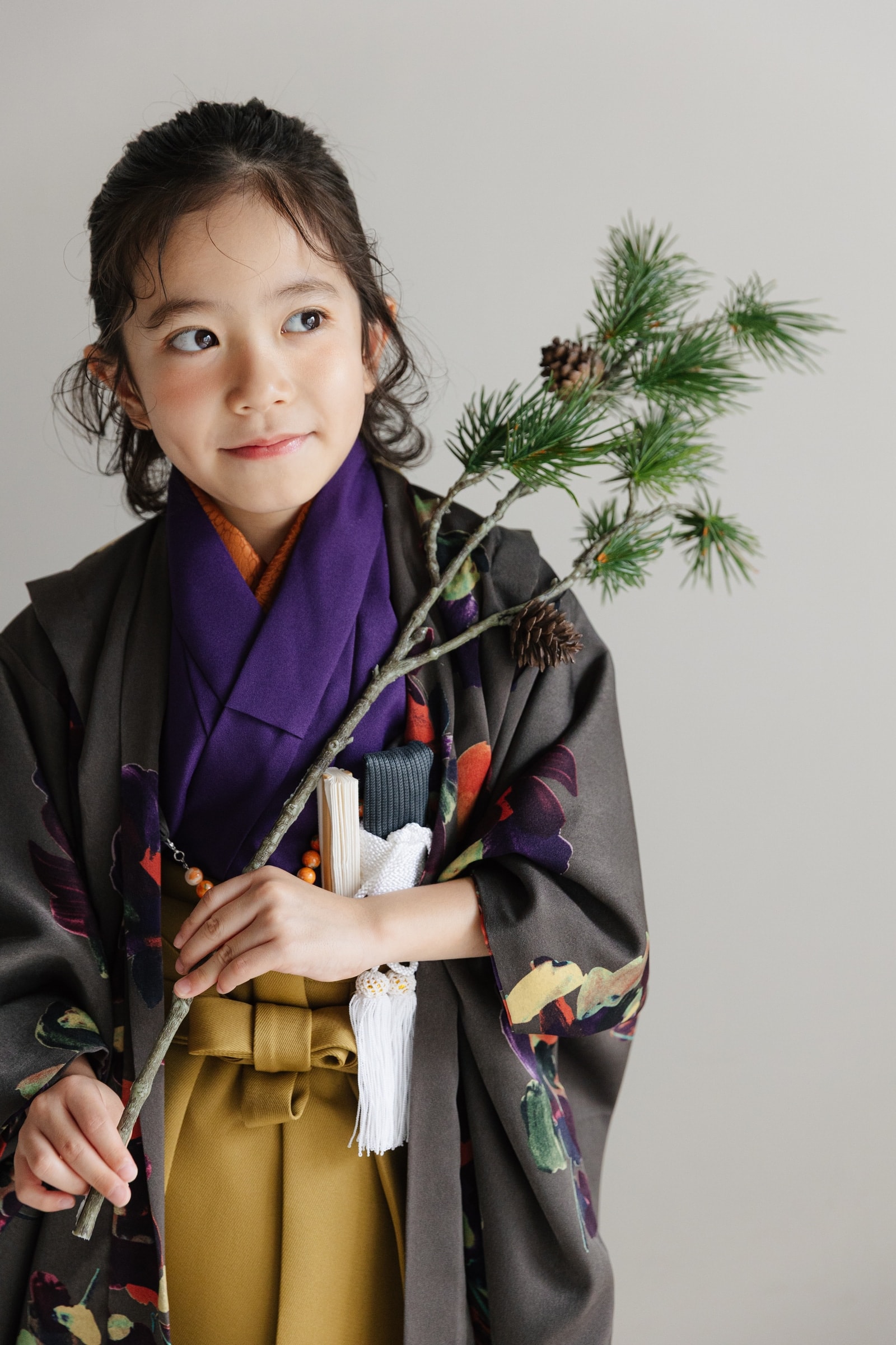 黒地に紫や橙の花柄を散らした羽織に紫の襟元、黄土色の袴を合わせた七五三の和装の子ども。松の枝を手に持ち、斜め上を見て微笑む上半身の構図。