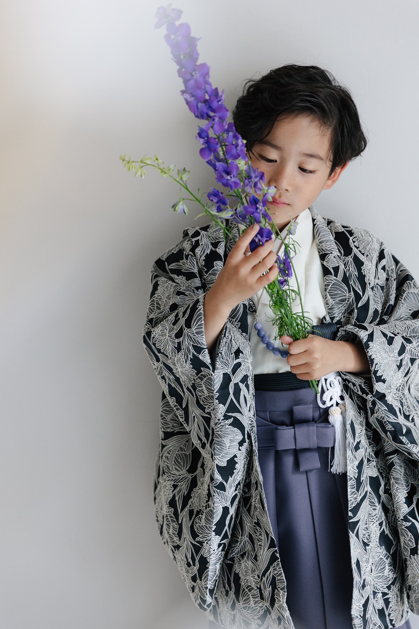 黒地に白の草花柄羽織と紫袴を着た男児が、紫色の花を手に持ち目を伏せた半身の構図。淡い灰色の背景で白い房飾りが下がる。
