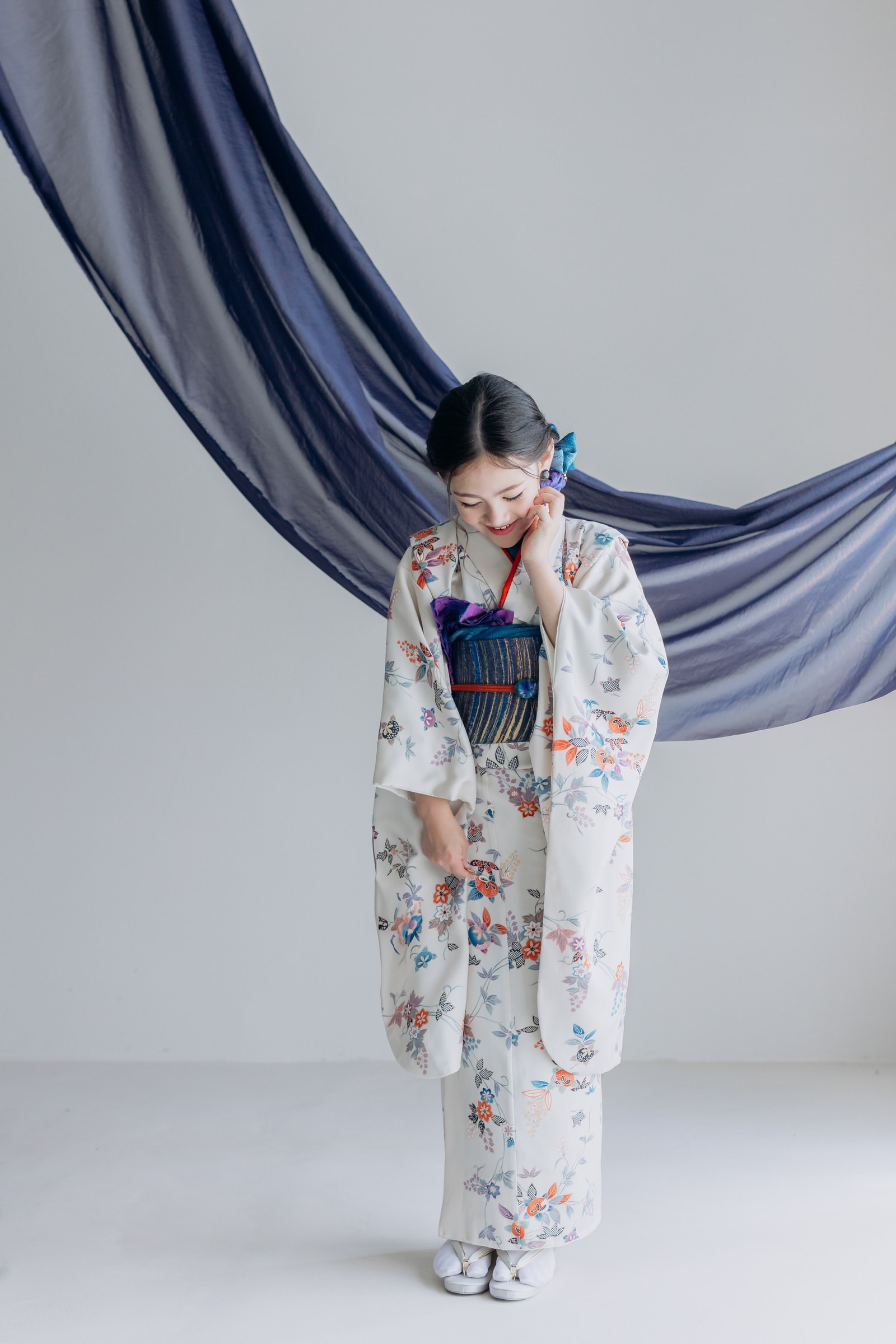 生成り地に色とりどりの花柄を散らした振袖を着た女性が、紺の縞帯を締めうつむき微笑む全身像。背景は白で藍色の薄布が弧を描く。