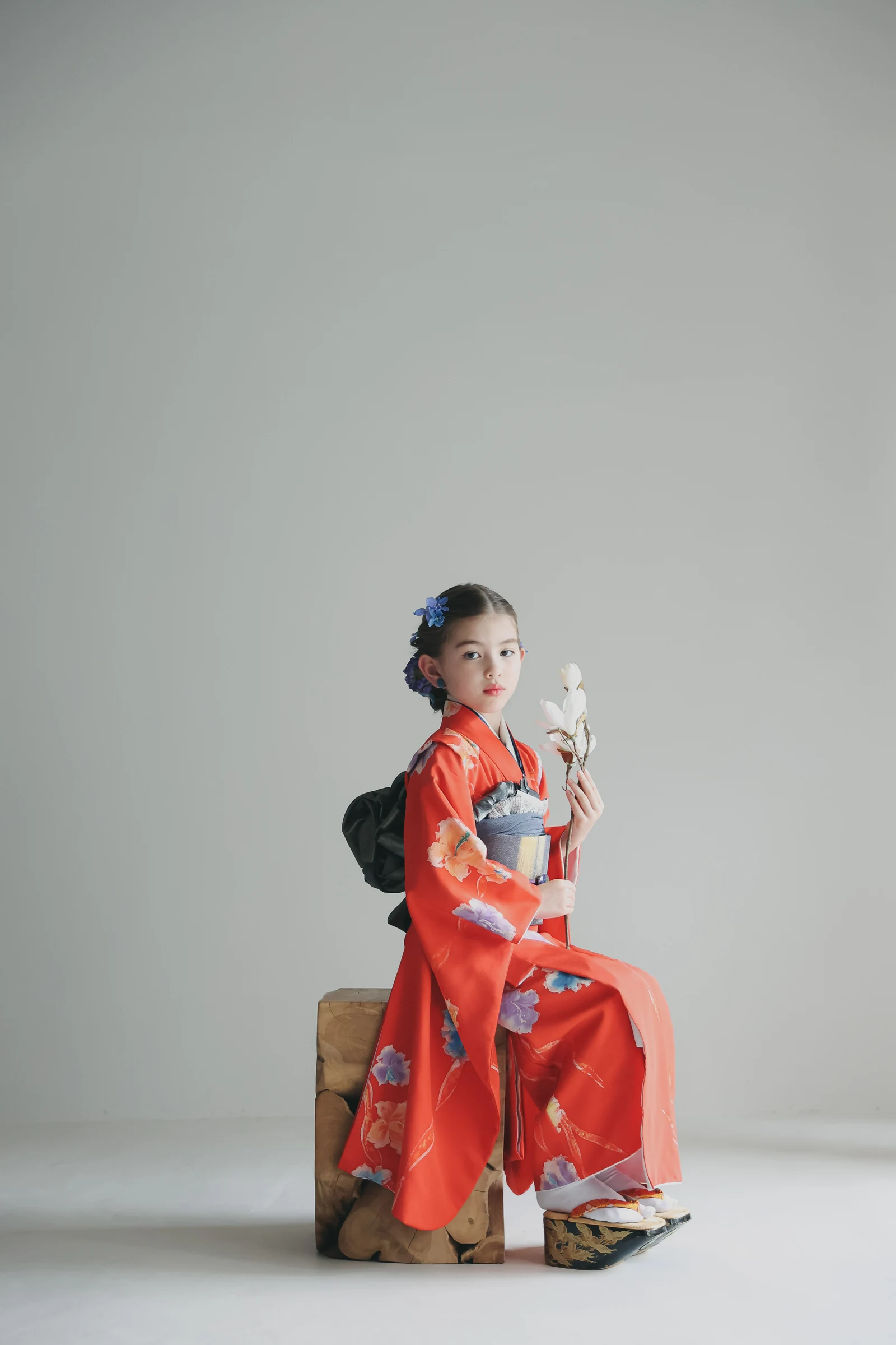 朱赤地に淡い紫やオレンジの花柄をあしらった七五三の着物姿の女児が、木の台に腰掛け、白い花の枝を手に横向きで座る全身構図。背景は淡いグレー。