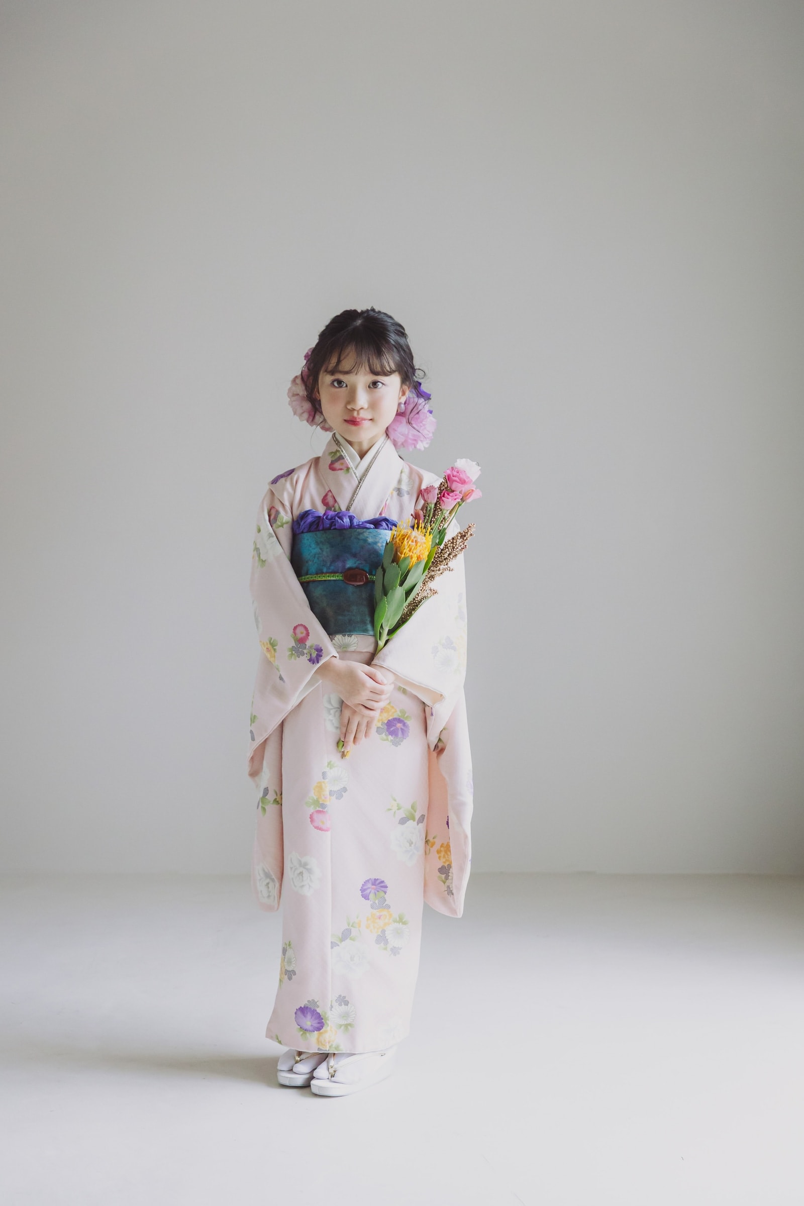 淡いピンク地に白や紫の花柄の着物を着た少女が、青緑の帯を締め、花束を抱えて正面に立つ全身像。背景は無地のグレーで白い足袋と草履を履いている。