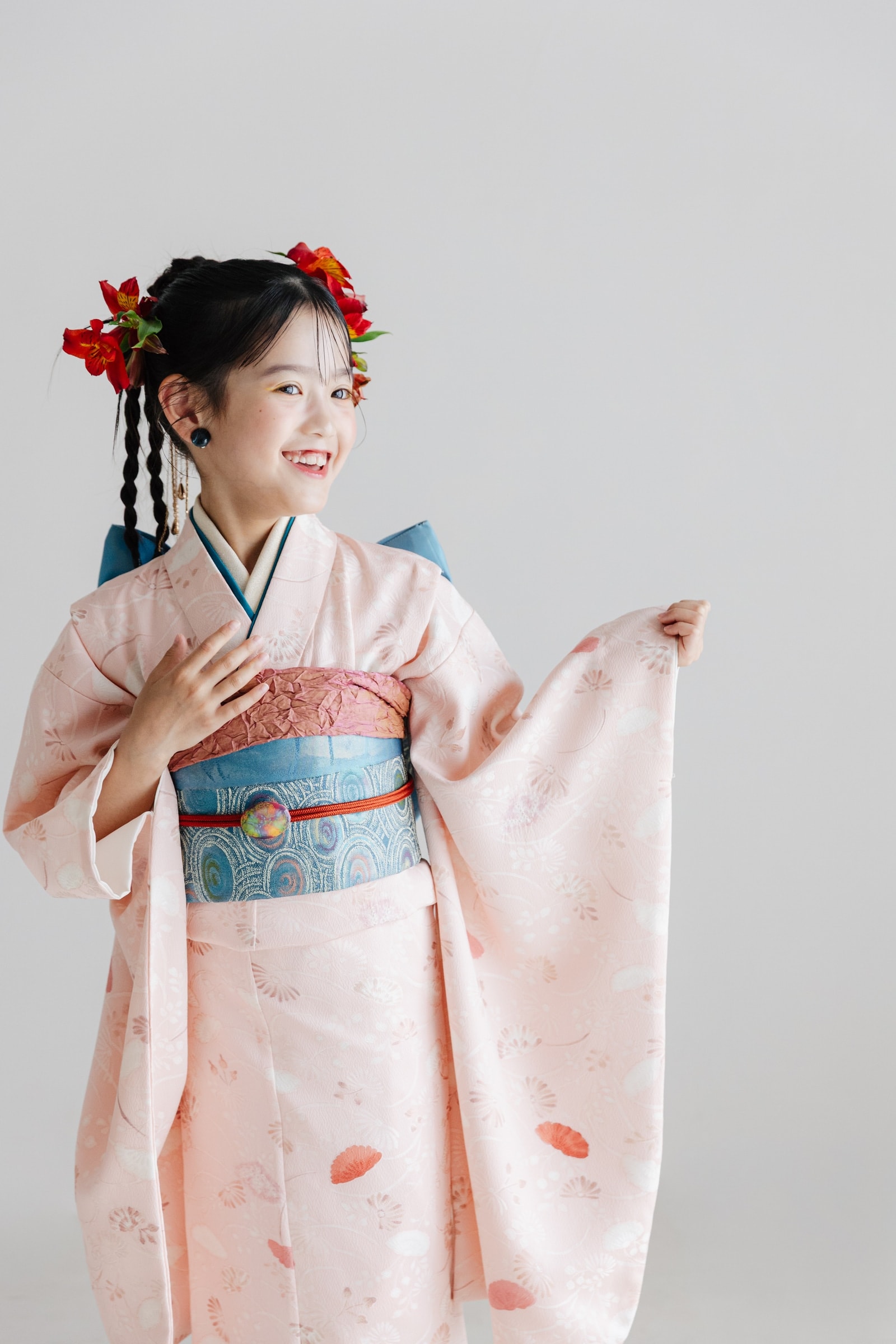 淡いピンク地に草花文様の着物を着た少女 — 青色の帯を締め、赤い花の髪飾りを付け、袖を広げて横を向き笑う。背景は明るいグレー。