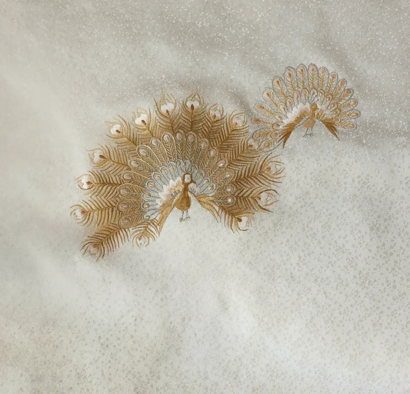 白銀地に金糸で羽を大きく広げた孔雀を二羽刺繍した七五三着物の生地クローズアップ。地には細かな銀の点描が散り、布の襞に沿って光沢が浮かぶ構図。