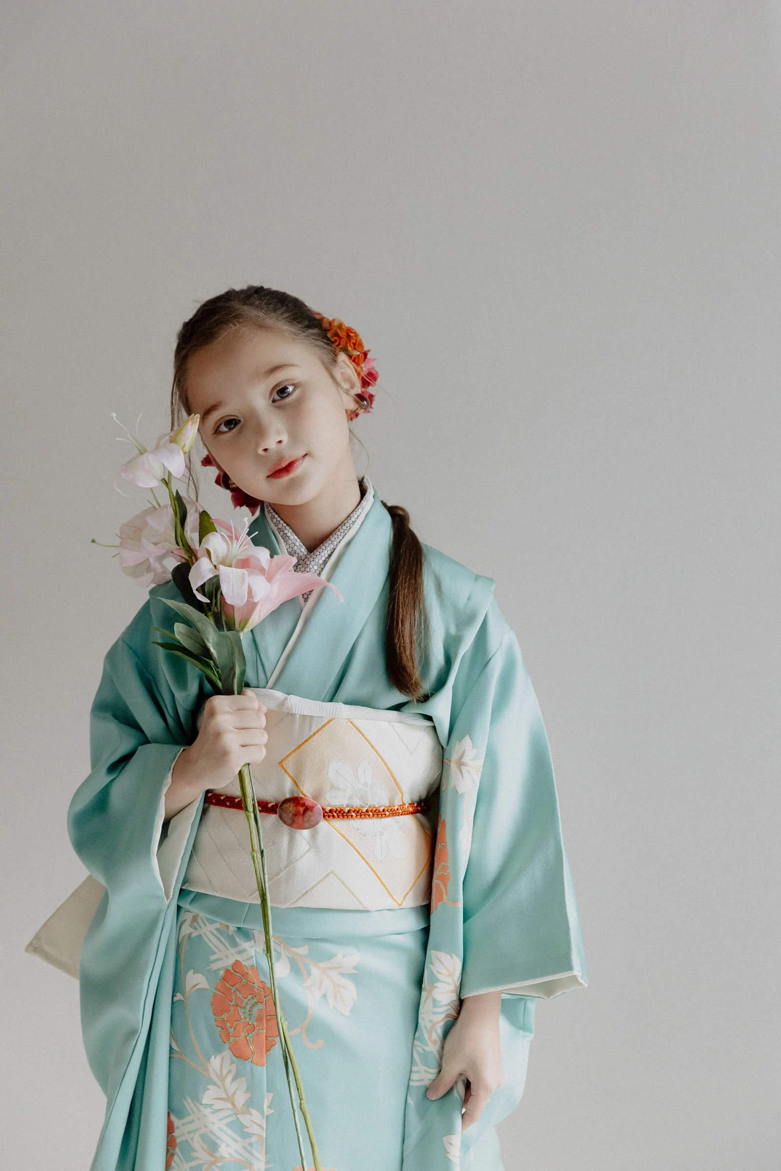水色地に橙と白の花柄を配した七五三の着物姿の女児。白い帯に赤い帯締め、髪に橙の花飾りを付け、ピンクの花を手に正面を向く半身構図。