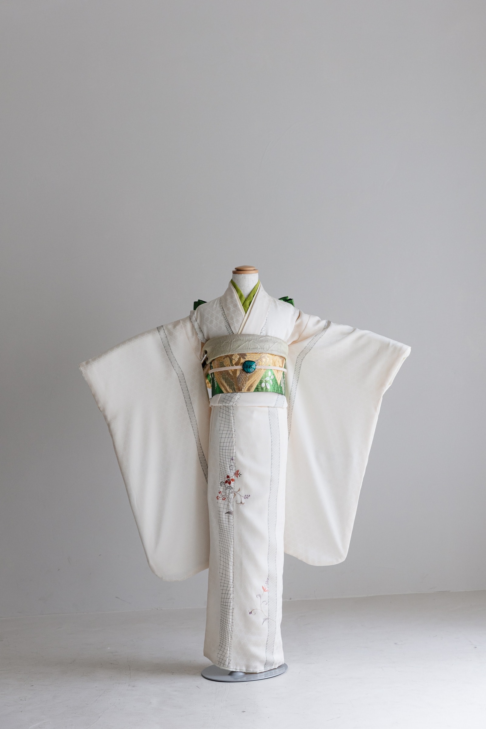 生成り色の振袖に格子と草花刺繍を配した着物 — トルソー正面、緑地に金彩の帯と緑の帯揚げを締め、両袖を広げた全身構図。