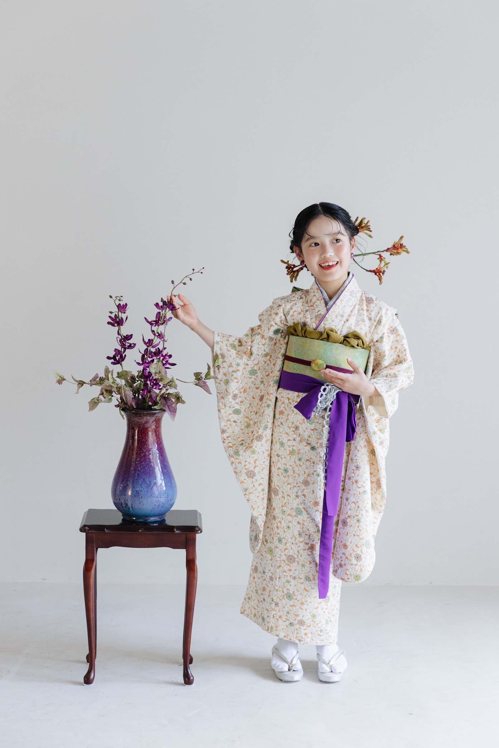 小花柄のクリーム色の着物に緑と紫の帯を締めた少女が、紫の花を活けた花瓶の横で枝に手を添えて立つ全身像。