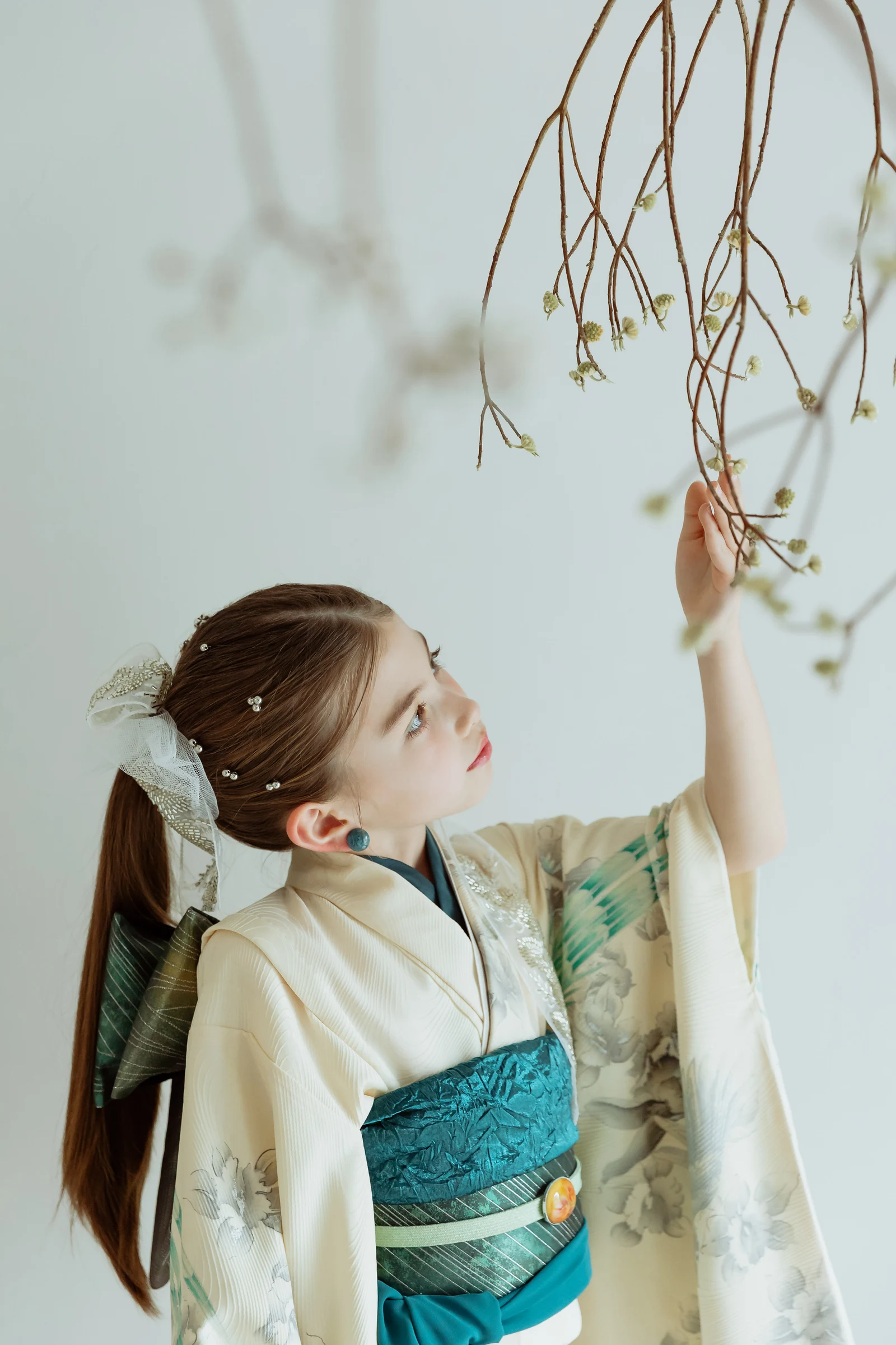 生成り地に淡い花柄をあしらった七五三の着物姿の女児が、青緑の帯を締め、片手を上げて頭上の枝に触れる横顔。明るい背景に長い髪と白いリボンが映る構図。