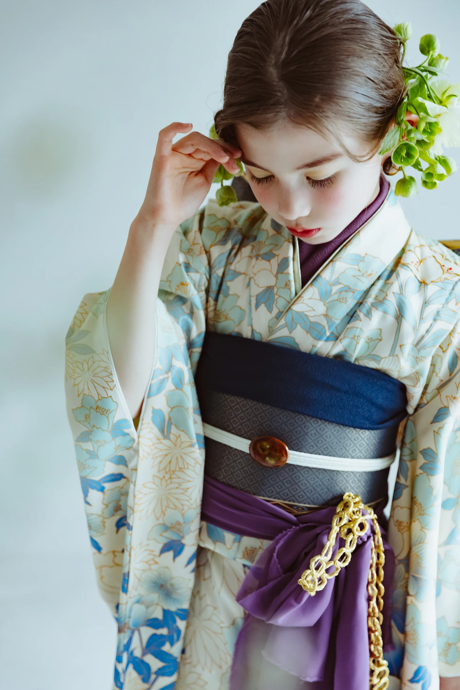 白地に水色の花柄の振袖を着た少女が伏し目で立つ半身像。紺と紫の帯に金の飾紐、髪に緑の花飾りを添え、片手を額に寄せる構図。
