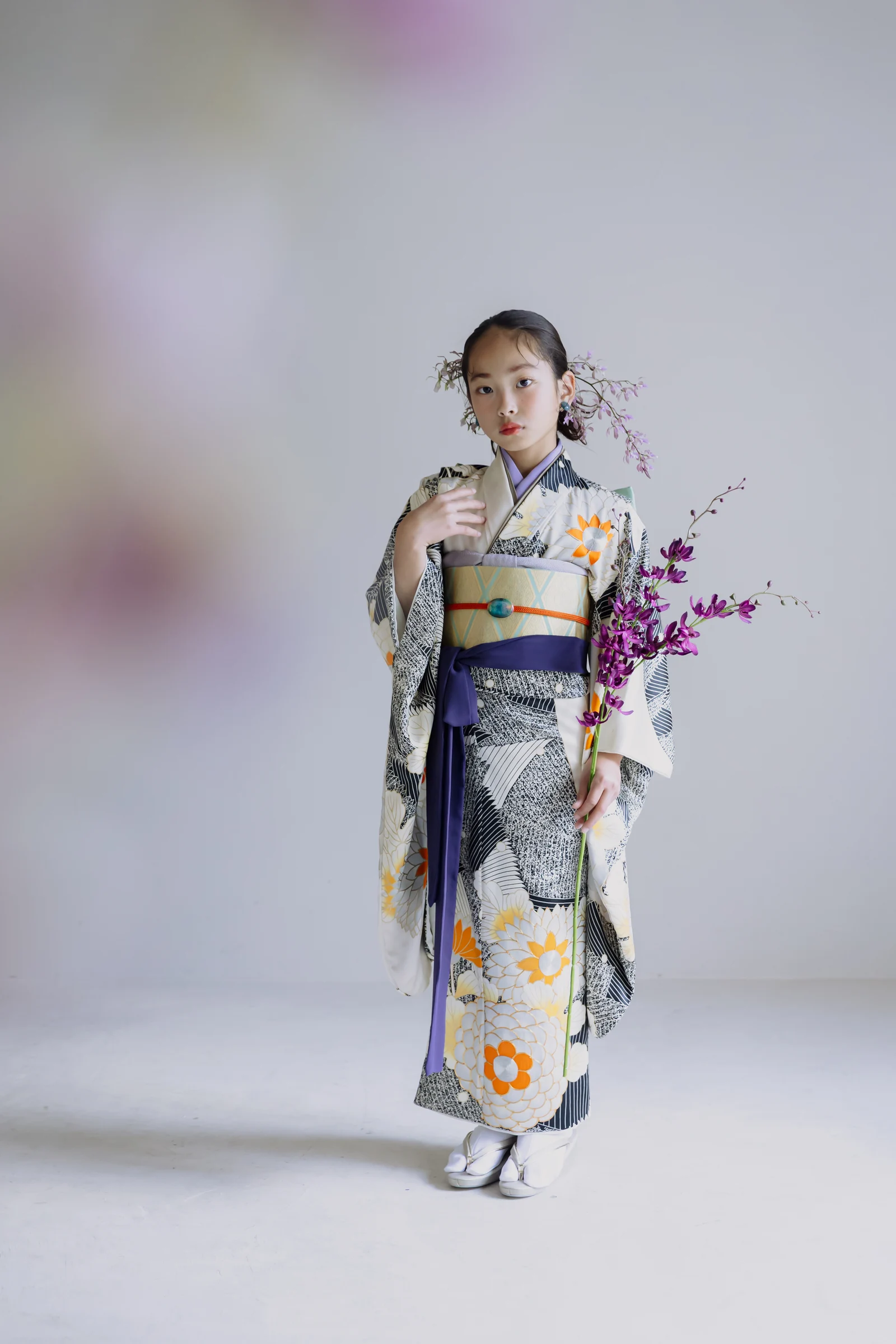 白地に黒と橙の大輪花柄の振袖を着た少女が紫色の蘭の枝を手に立つ全身像。黄の帯に紫の帯締めを合わせ、淡い背景の正面構図。