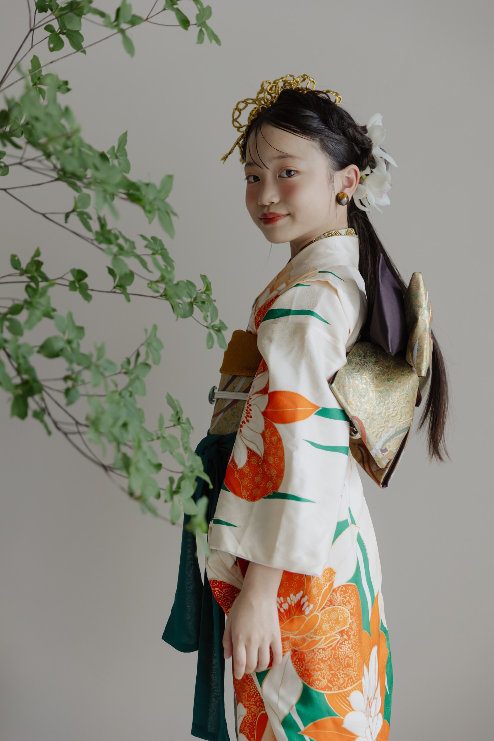白地にオレンジと緑の大きな花柄が描かれた振袖姿の少女 — 金の髪飾りと白い花を挿し、後ろ姿から振り返る半身構図。左手前に緑の枝葉。