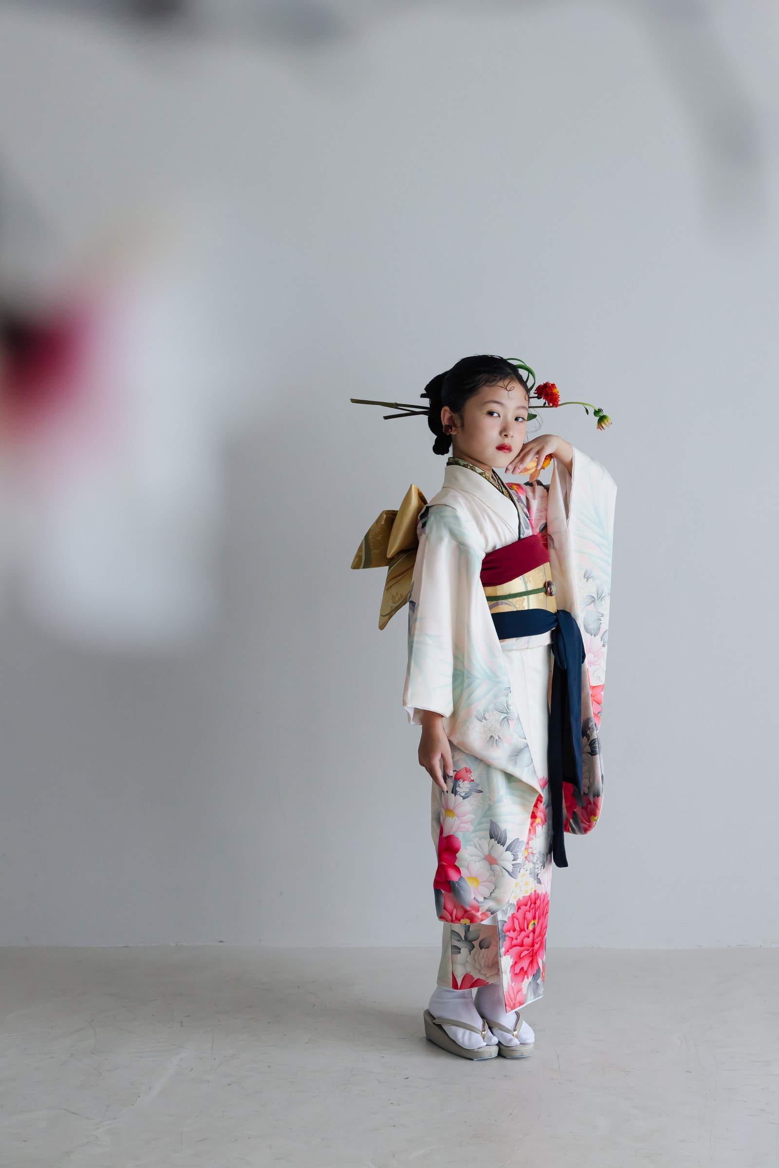 白地に赤や桃色の花々を描いた振袖に金の帯を締めた少女 — 髪飾りを付け、造花を手に横向きで立つ全身像、背景は淡いグレー。
