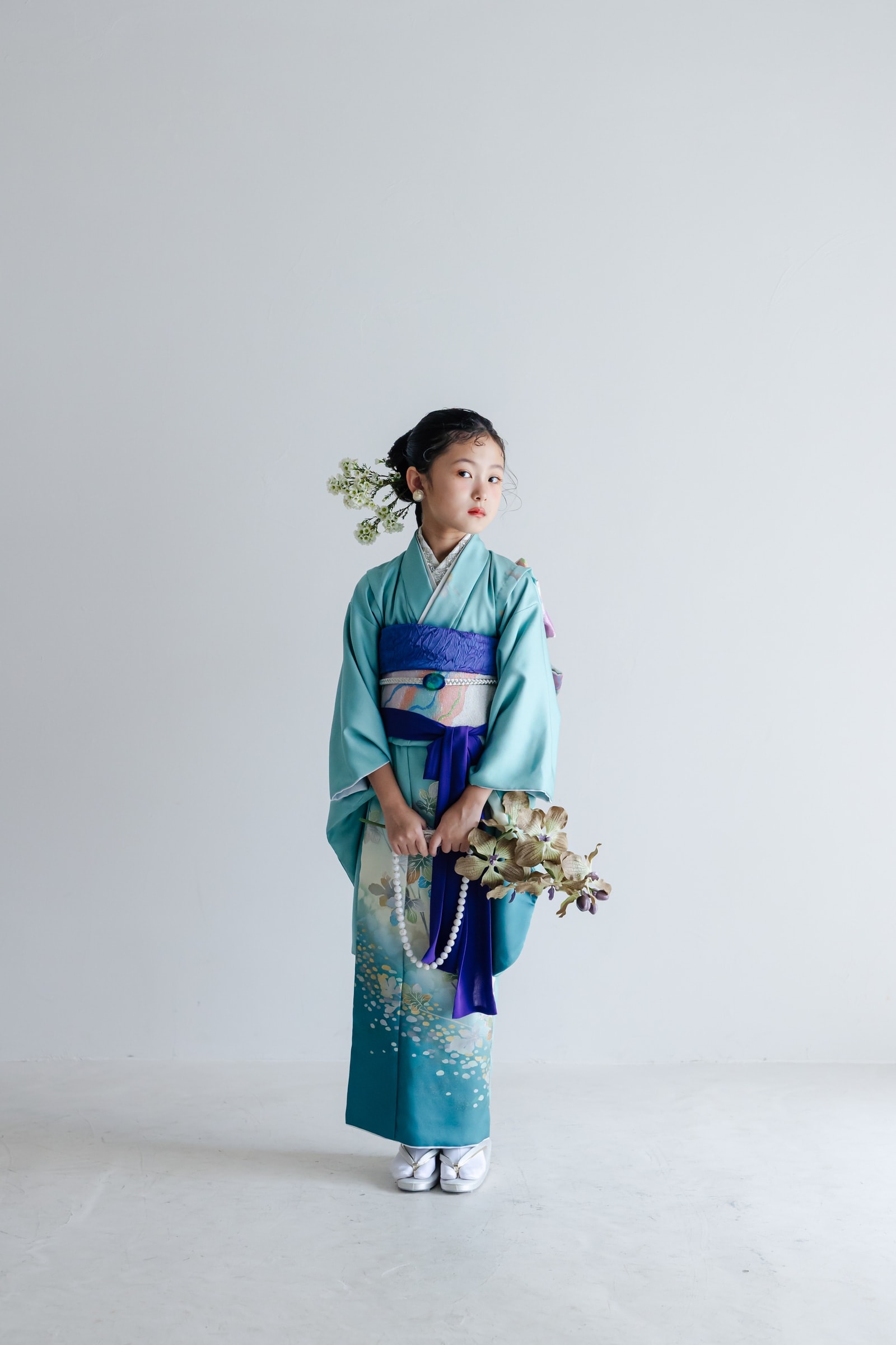 裾に向かって濃くなる翠色の花柄着物を着た女児の全身像。紫の帯揚げと白い帯締めを合わせ、髪に白い花飾りを付け花を手に正面を向いて立つ。