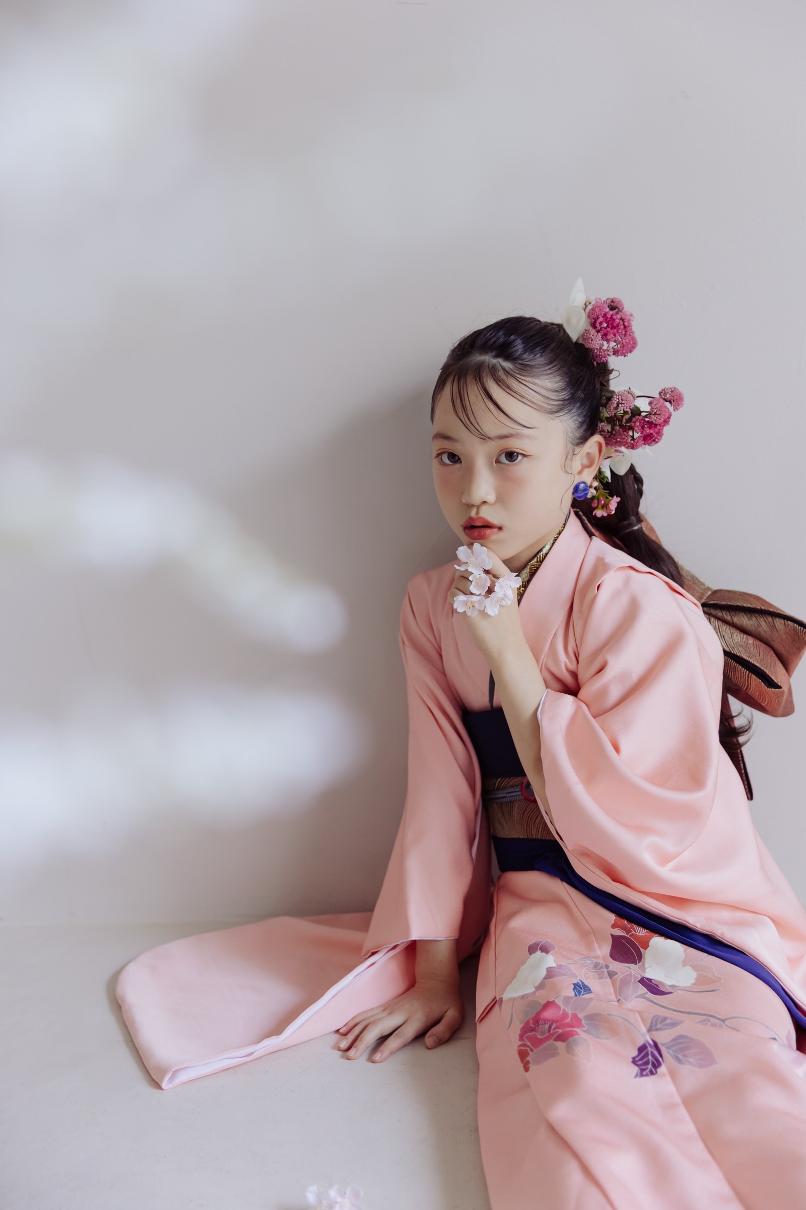 淡い桃色地に花柄と紺の帯を合わせた着物姿の少女が床に座り、髪に紅白の花飾りを付け、桜の花を手に持つ。淡色背景の縦構図。